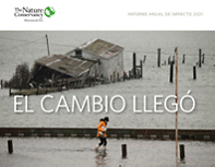 Cover of the Spanish translation of the 2021 MD/DC Impact Report. A woman wearing a yellow rain slicker and tall rubber boots walks through ankle deep water on a flooded street. 