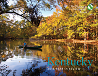 A person kayaks on a placid body of water in a wooded area.