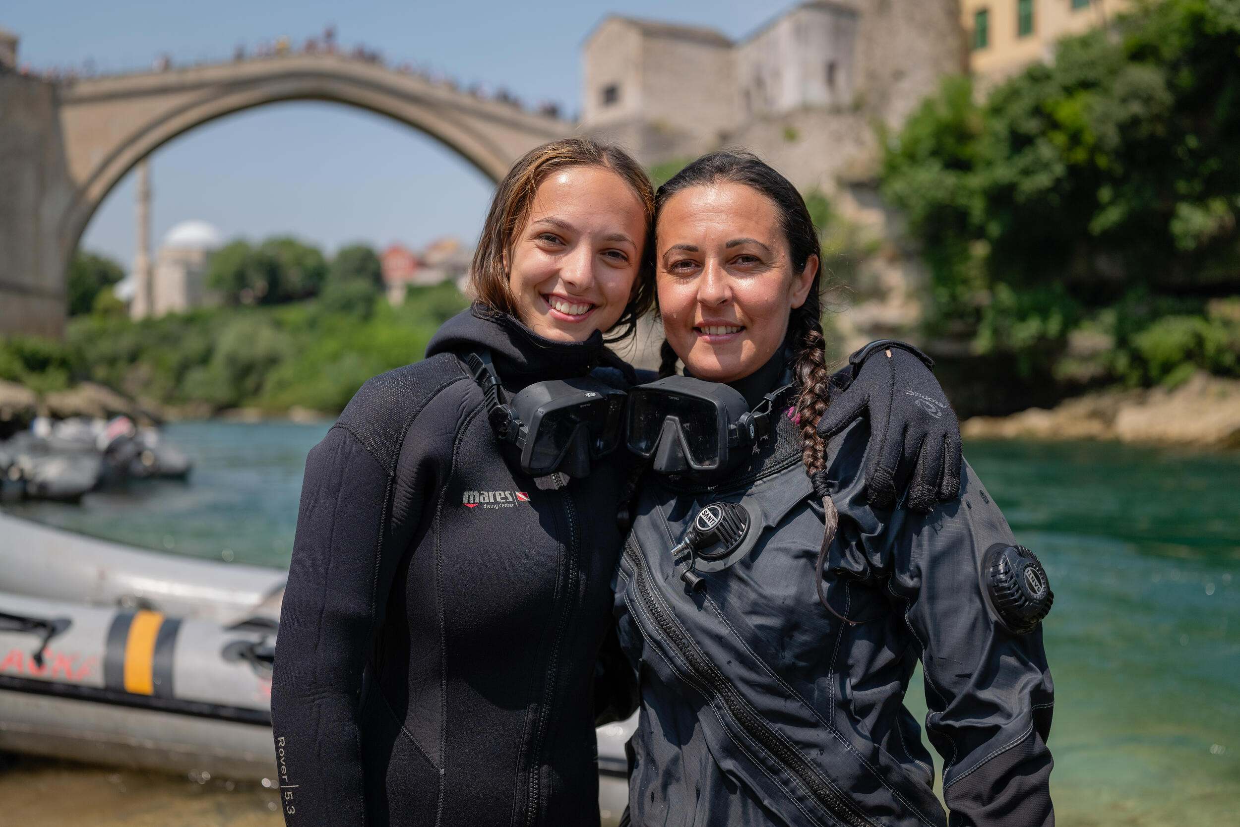 Two women wearing black drysuits pose smiling with a greenish blue river and grey and yellow raft behind them. A distant stone bridge behind the women crosses into a town on a hill.