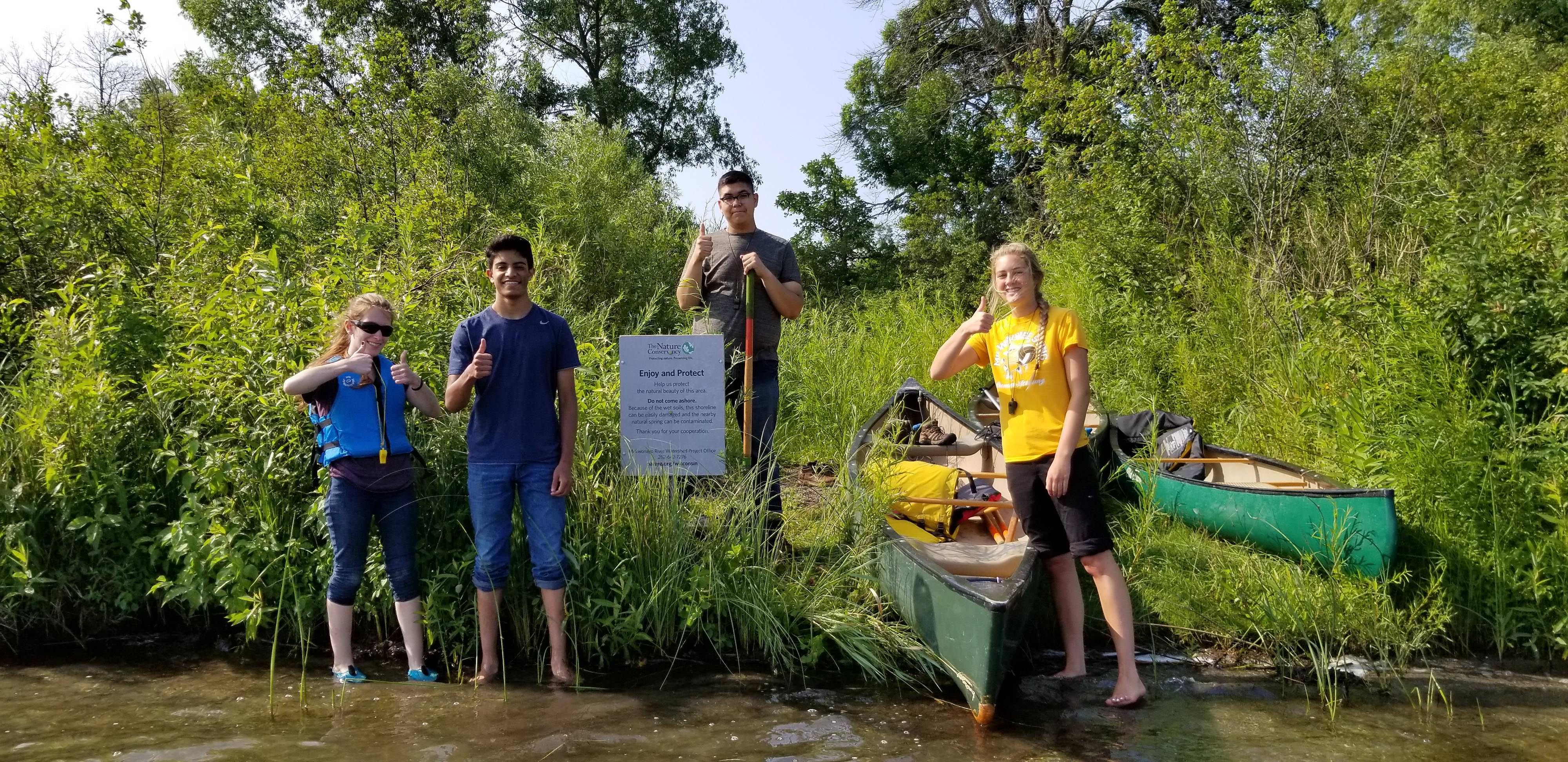 Teenagers posing in front of canoes at a lake. 