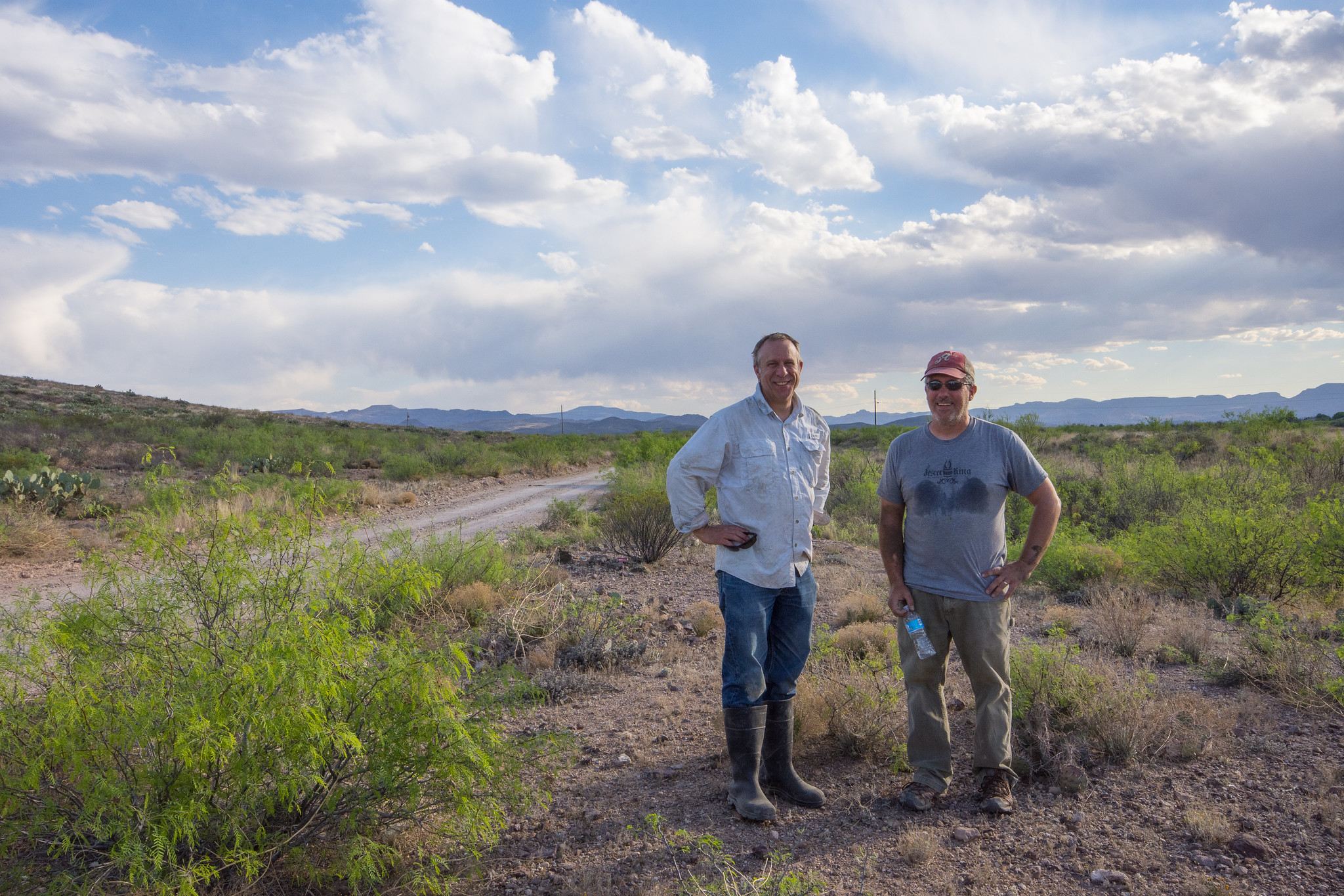 Two men stand next to each other near a desert road.