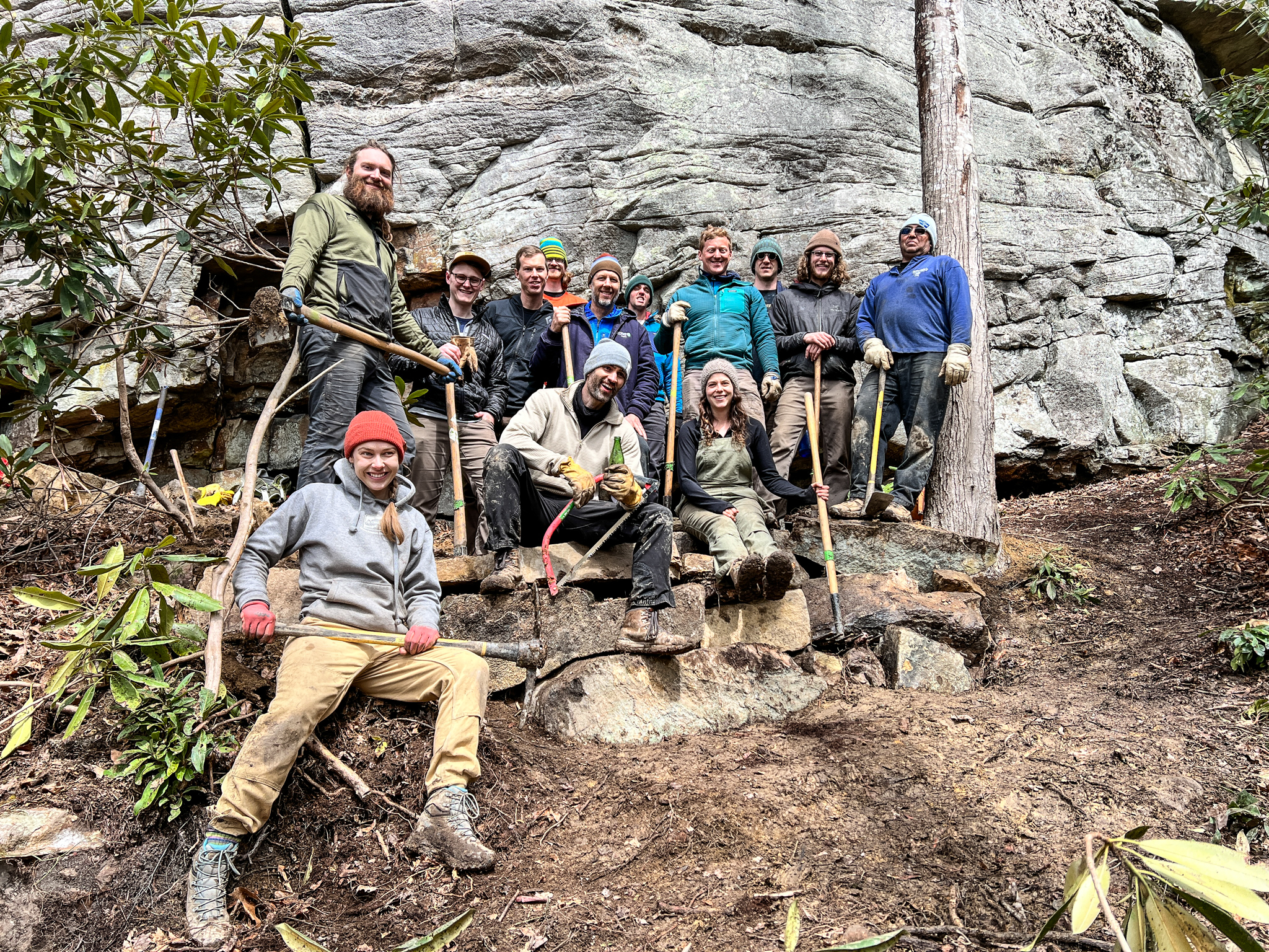 A large group of people pose together in front of a rock wall. They hold loppers and other hand tools for clearing brush and small vegetation.