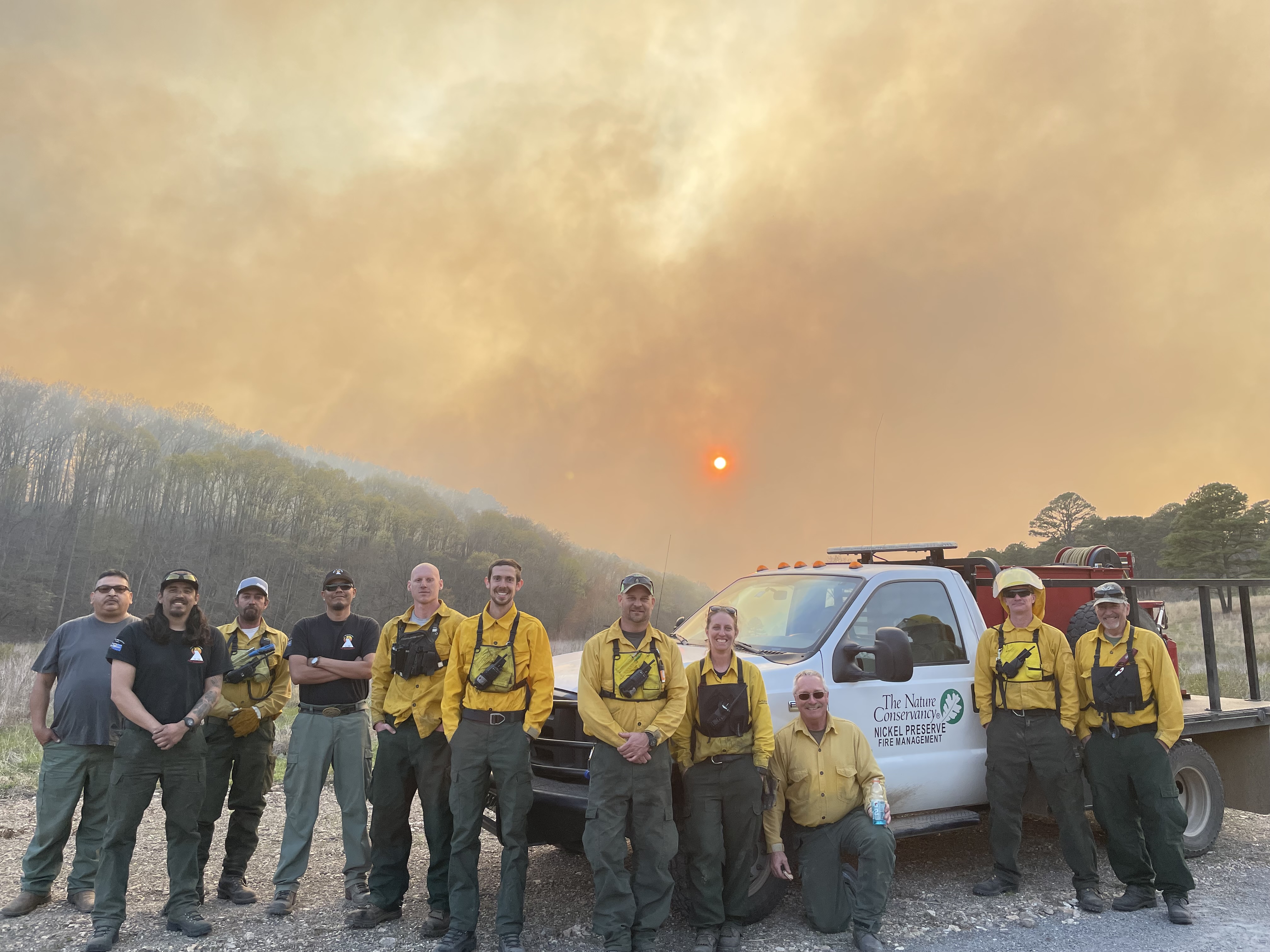 Fire crews of the Cheyenne and Arapaho Tribes and The Nature Conservancy in Oklahoma pose for a photo in front of a smoky sunset. 
