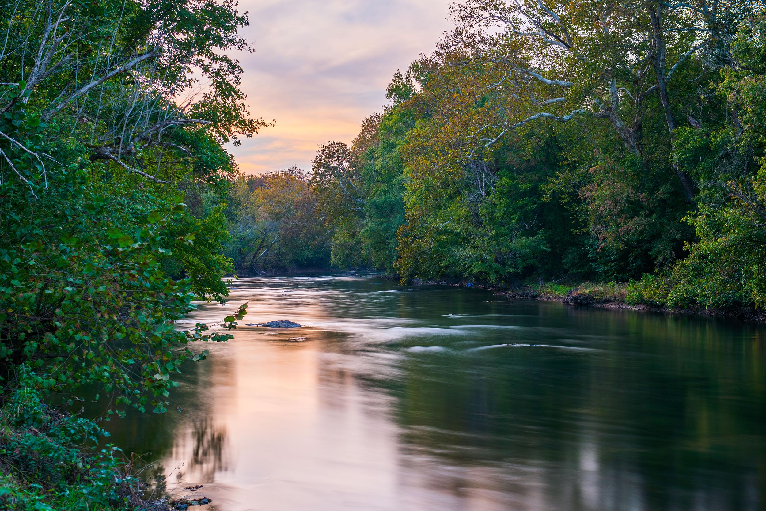 A wide, flat river flow between tree lined banks.