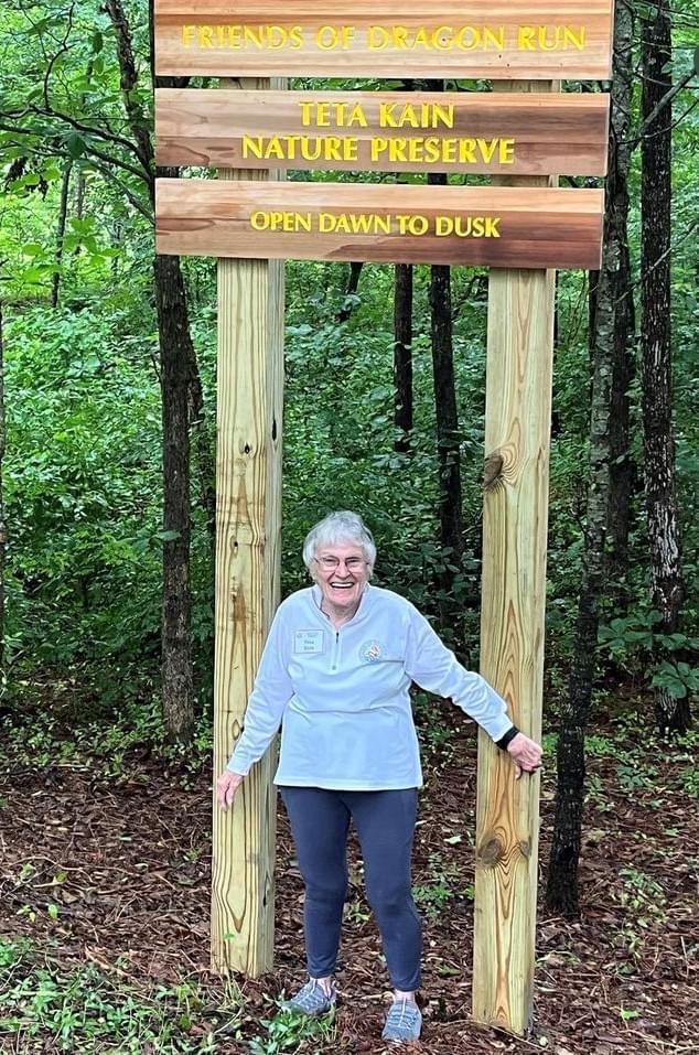 A smiling woman stands underneath a tall preserve sign that reads, Friends of Dragon Run Teta Kain Nature Preserve, Open Dawn to Dusk.