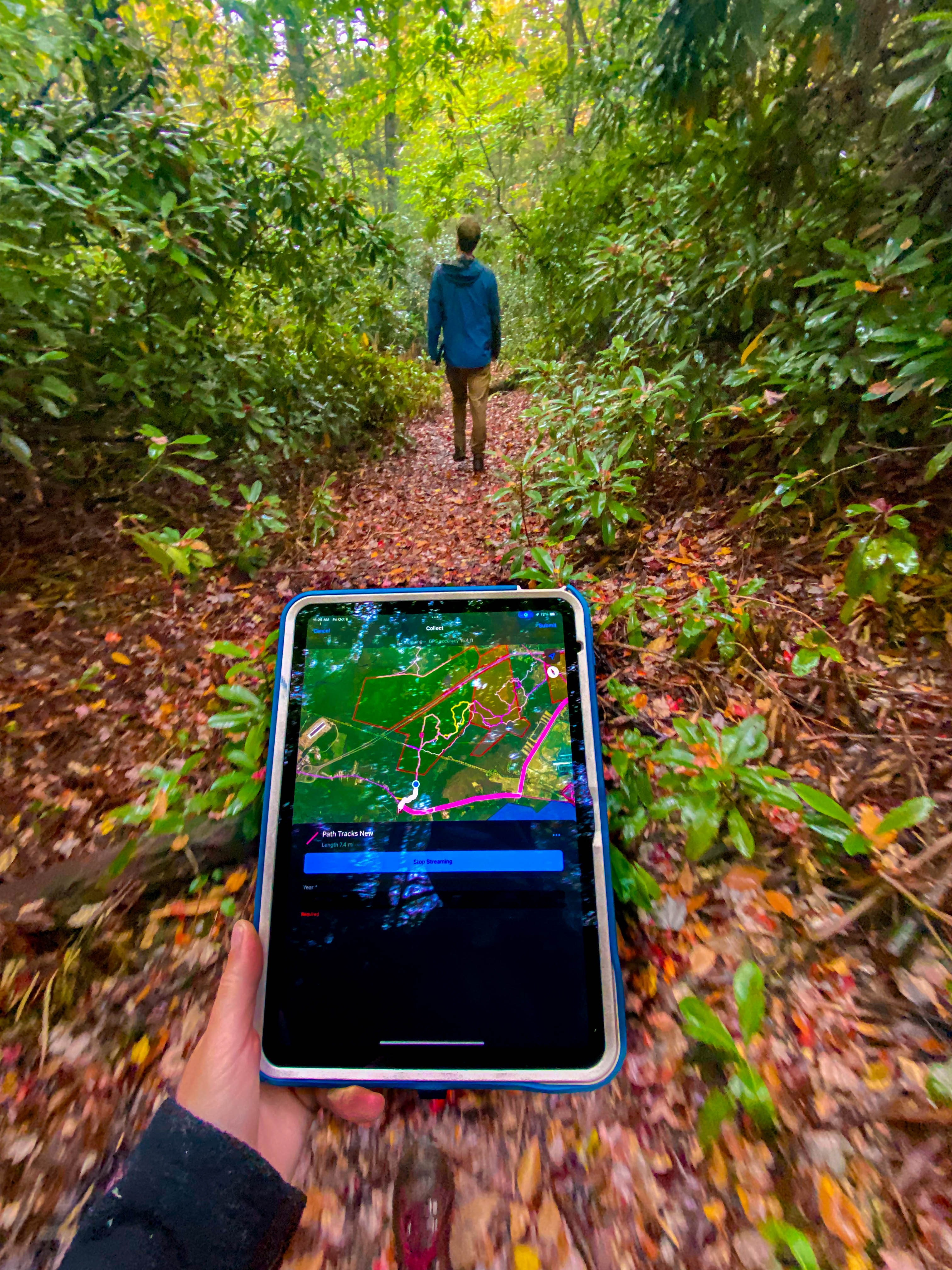 A hand holds a tablet with a view of a map in front of them while walking along a trail littered with orange leaves.
