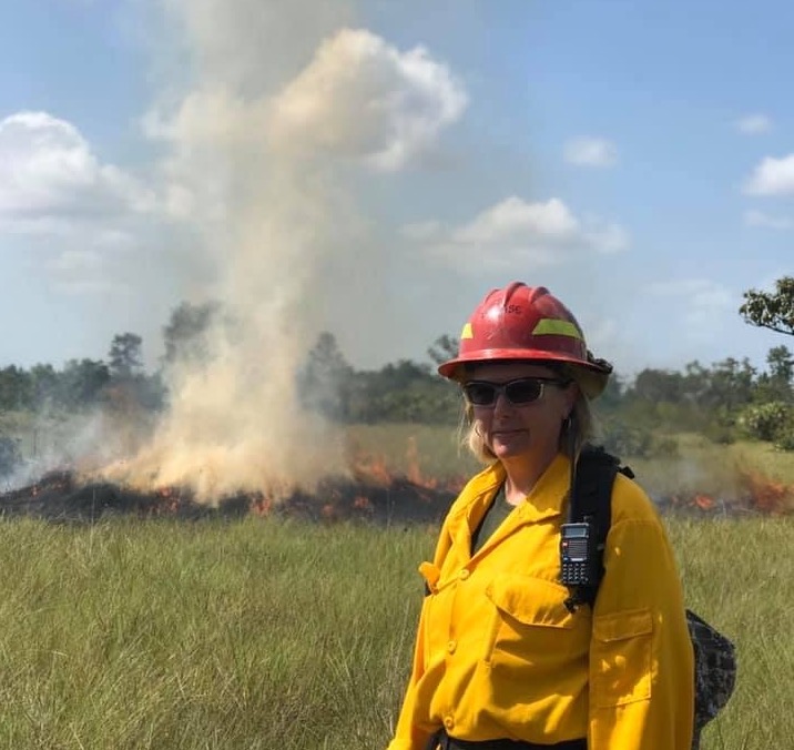 A photo of Jenny Case in fire gear standing in front of a small smoky fire in a field.