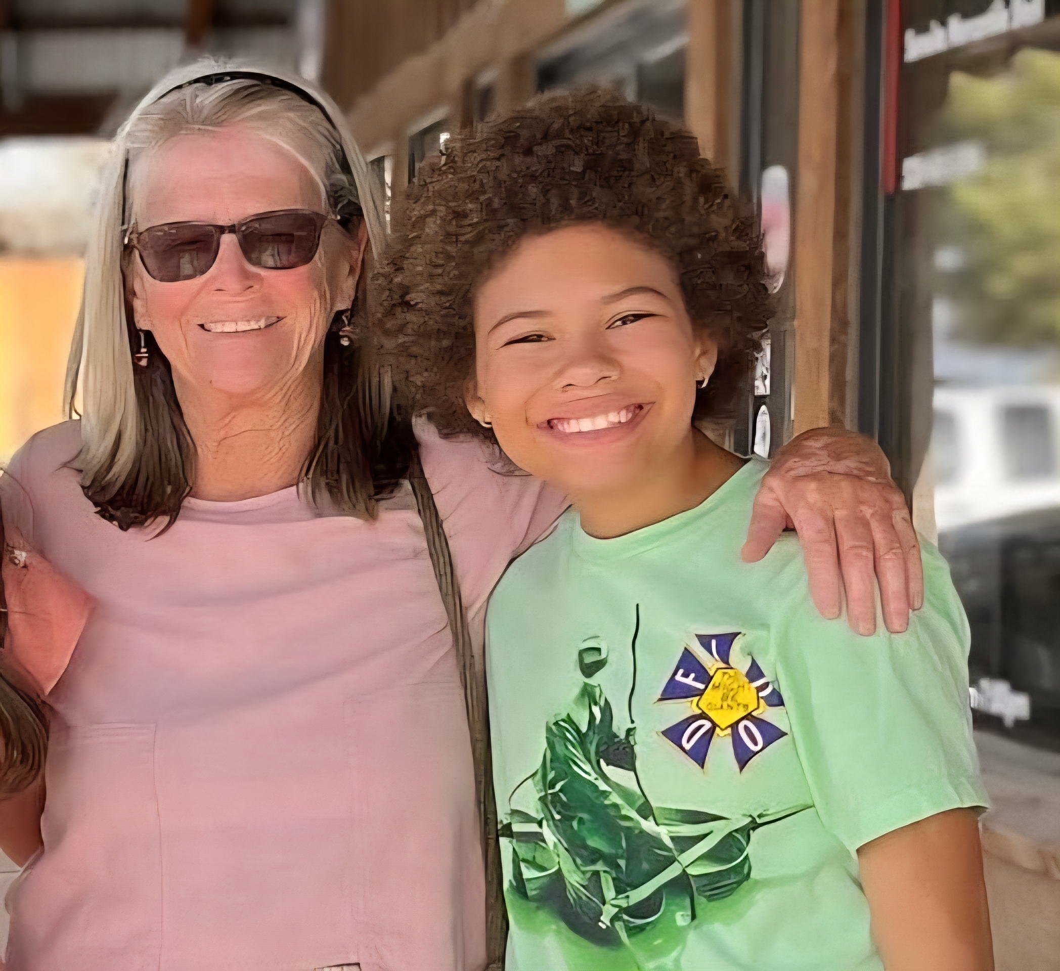 A woman wearing sunglasses poses with her arm around a young person.