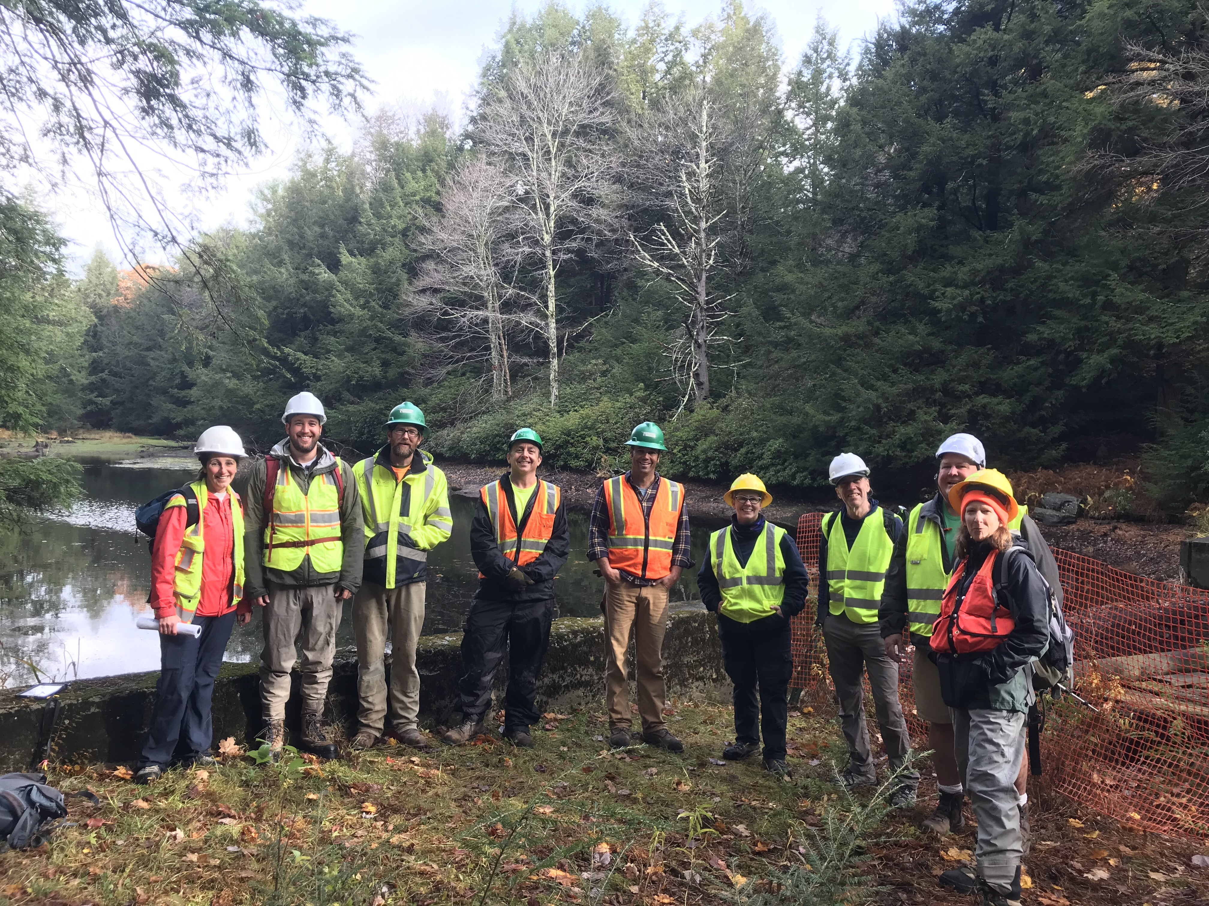 A group photo of nine people in orange and yellow safety vests and hard hats standing at Becker Pond in Mount Washington, Massachusetts. 