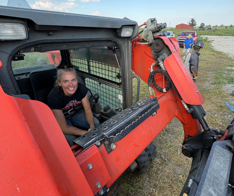 Olivia Schouten sits smiling in the cab of a compact piece of construction equipment outdoors, with one hand near the controls.