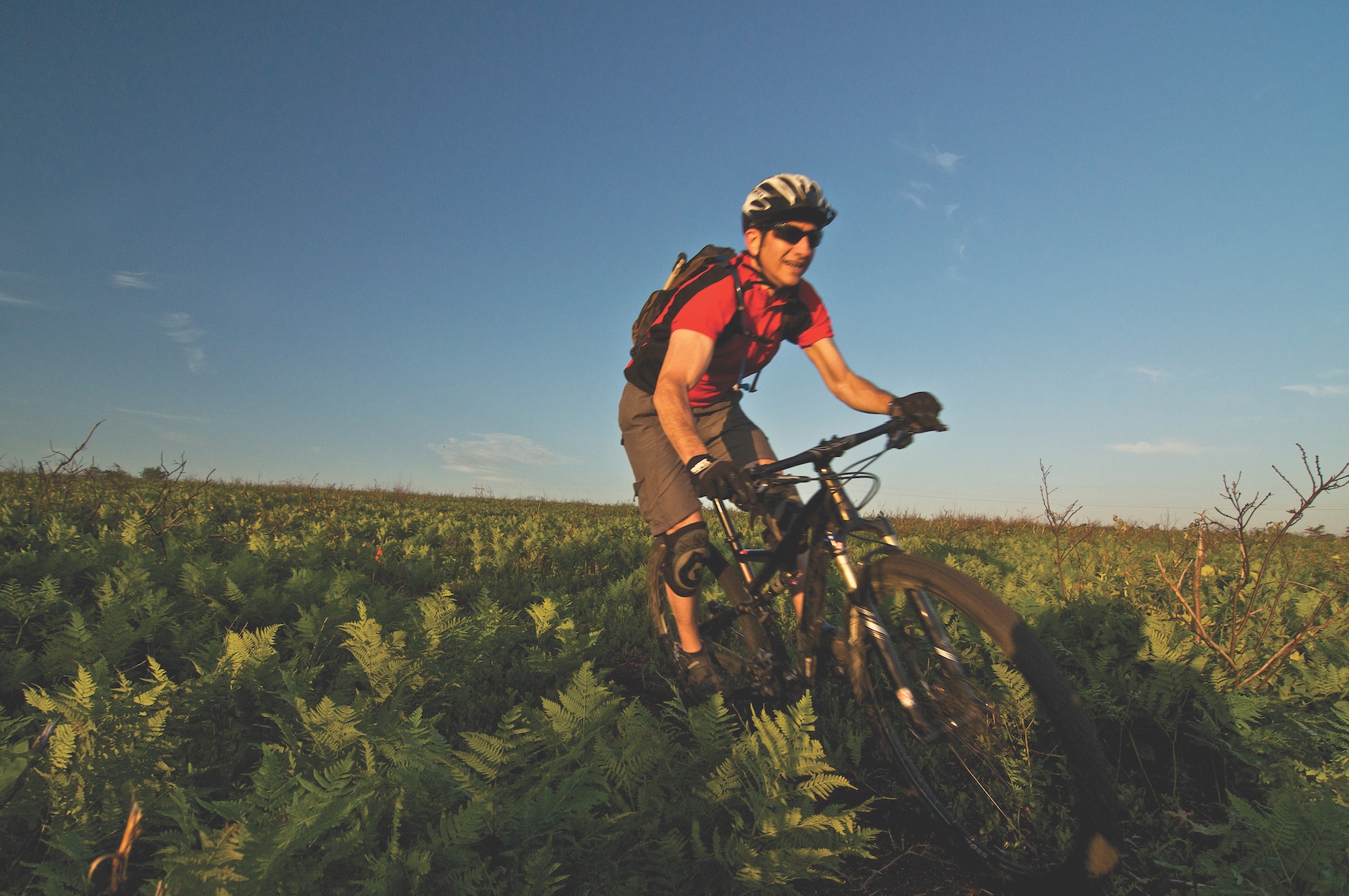 A bike rider wearing a red shirt and helmet moves across a grassy field.