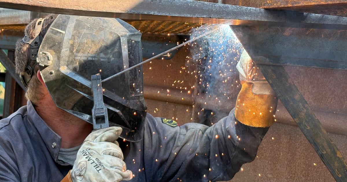 A man wearing protective welding gear works on a metal section of the bison corrals as bright sparks fly from the welding point.