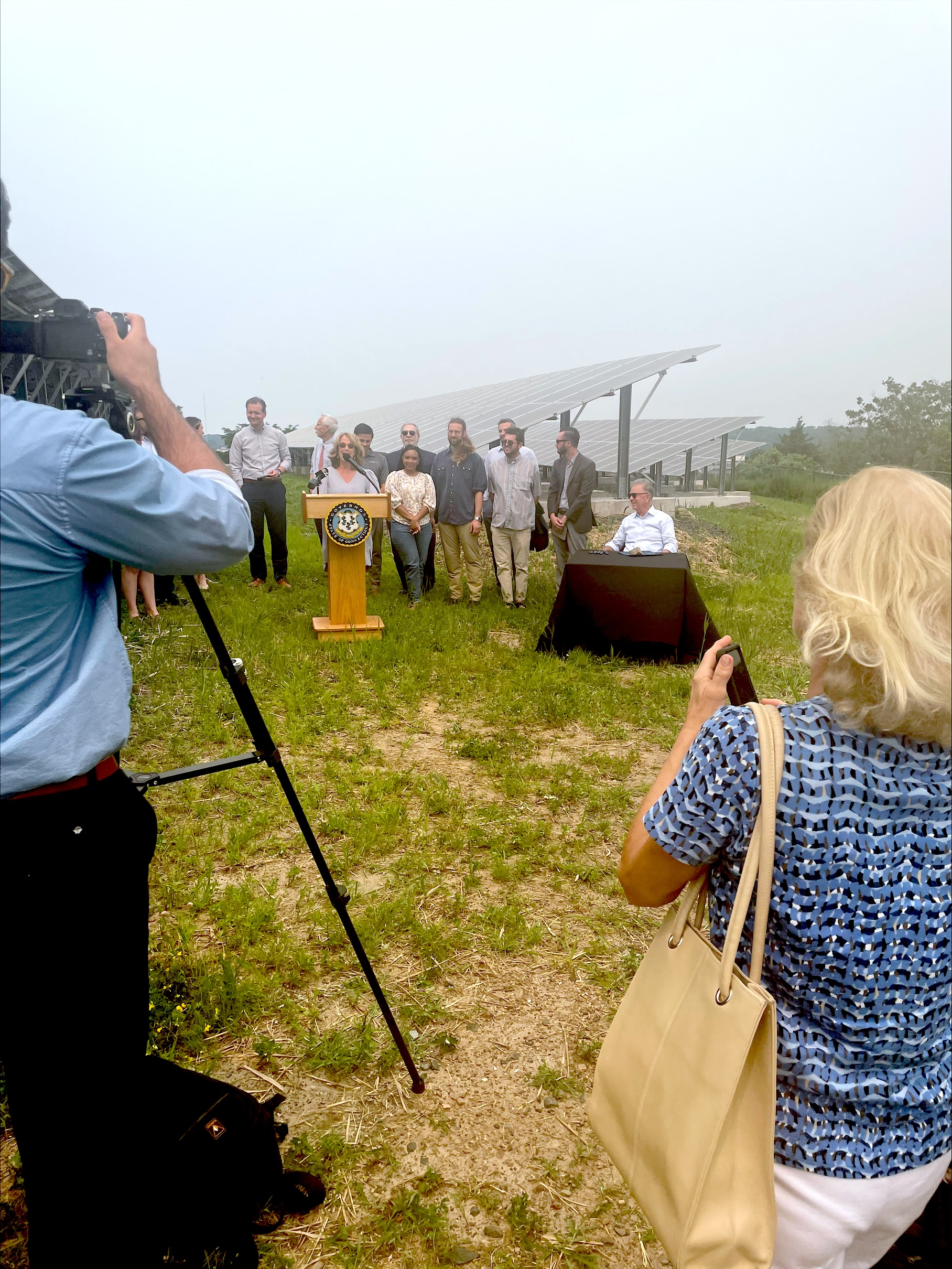 A group of people stands outdoors near a podium with an official seal, surrounded by solar panel installations. One individual is seated at a table with documents. People are taking photos and videos of the scene. The background includes greenery and more solar panels.