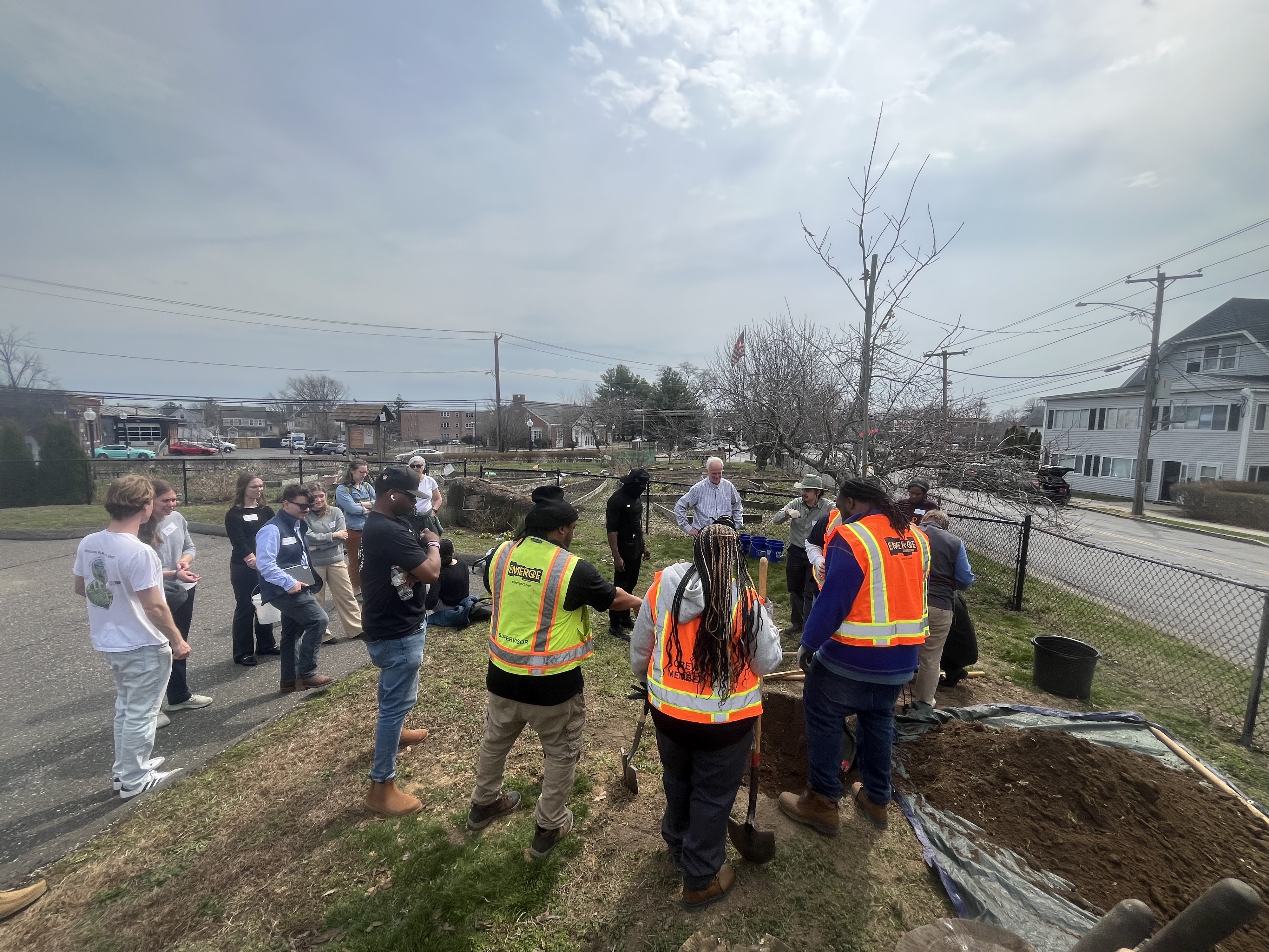 A group of individuals gathers outdoors near a tree with freshly dug soil and a tarp. Several people wear fluorescent safety vests with "Verizon" written on them. A few people sit on the grass, while others stand nearby. Residential houses and power lines are in the background under a cloudy sky.