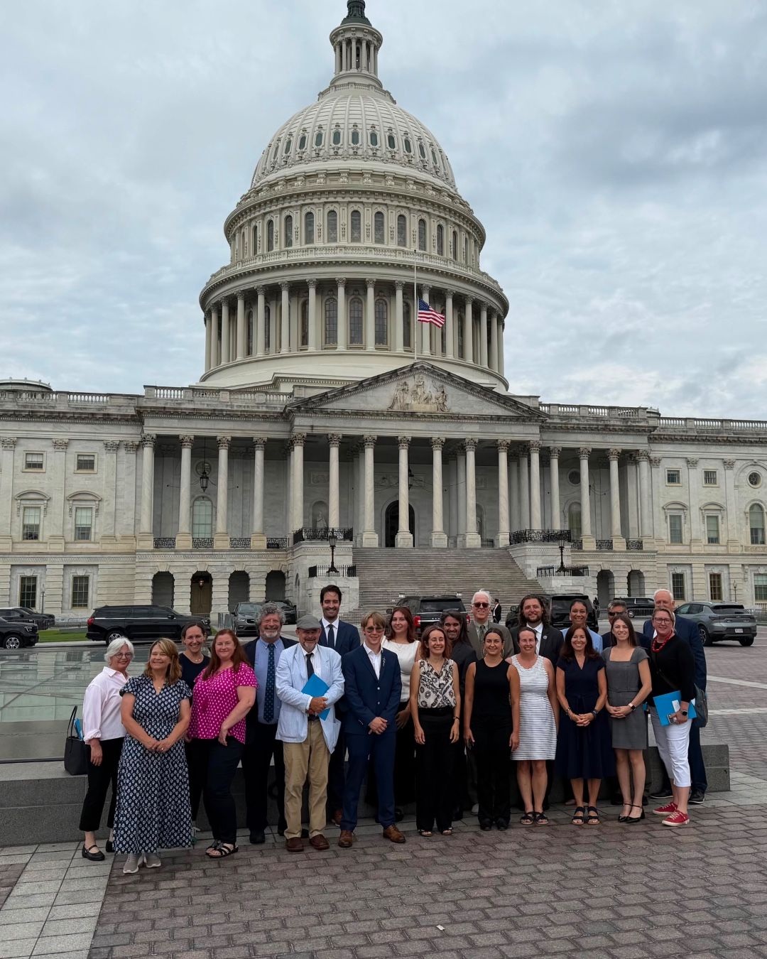 A group of people stands in front of the United States Capitol building on a cloudy day. The Capitol's dome and columns are prominently displayed in the background.