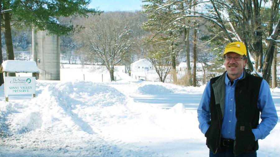 A person stands in a snowy landscape near a sign that reads "Sunny Valley Preserve The Nature Conservancy." They are wearing a yellow cap, glasses, and a dark vest over a blue shirt. Trees and a silo are visible in the background.