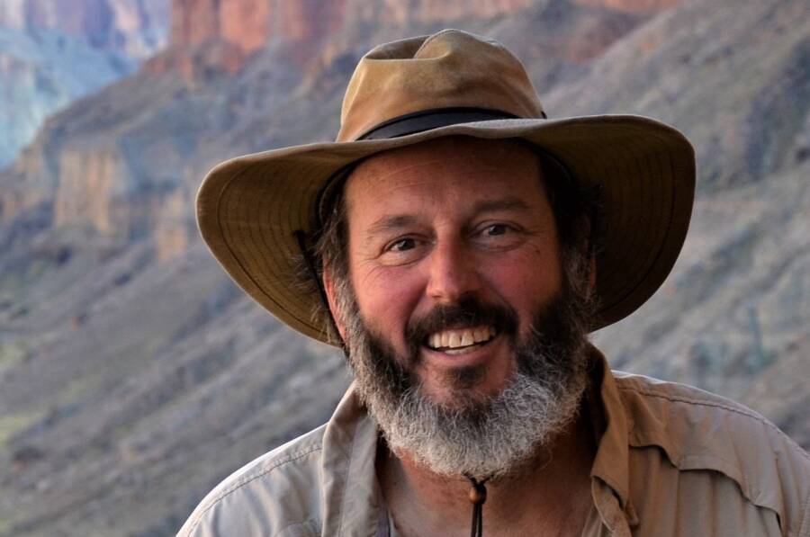 A person outdoors wearing a wide-brimmed hat and a shirt, smiling with a rocky landscape in the background.