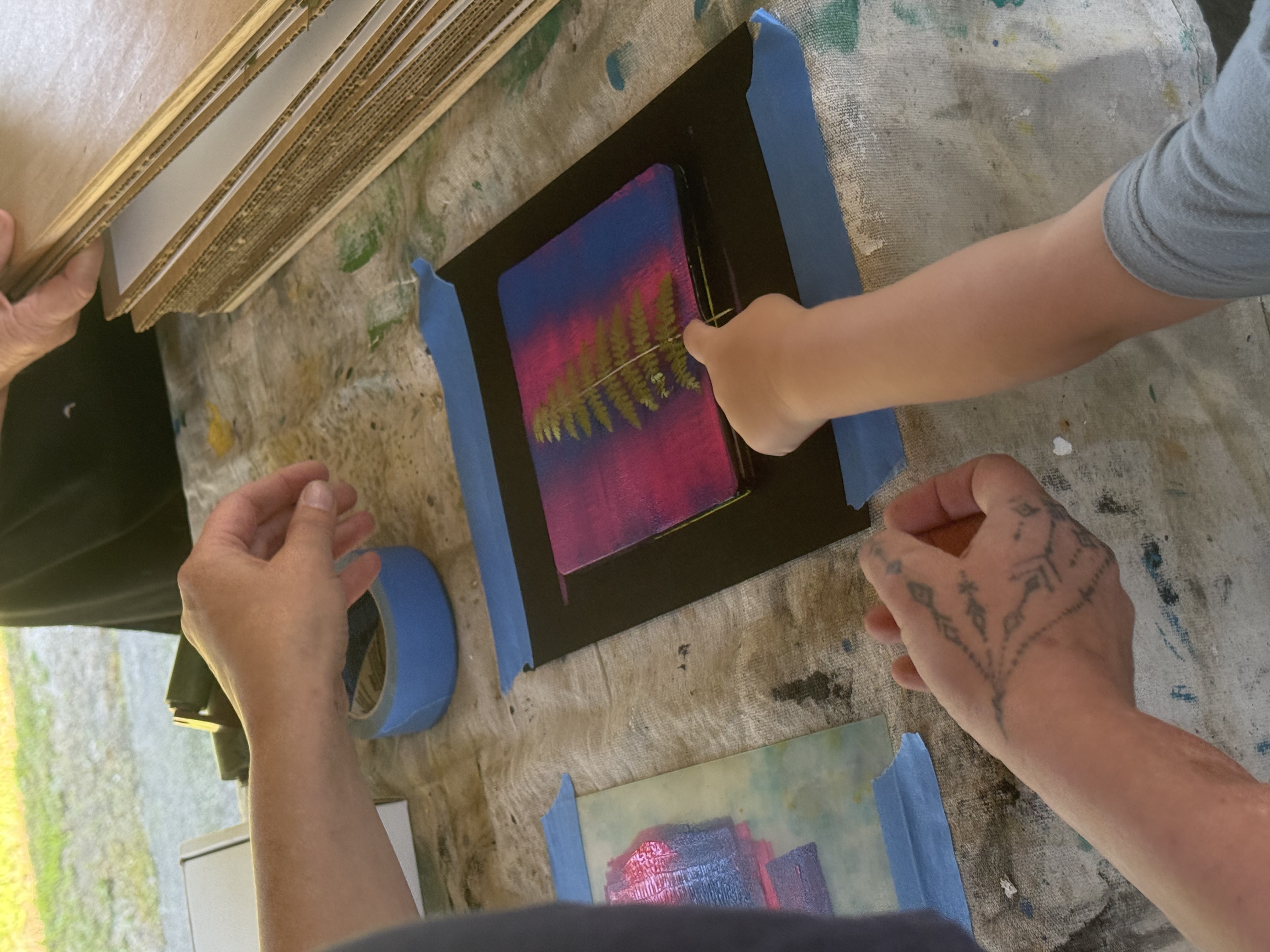 Two individuals are engaged in an art activity, applying paint onto a small canvas that features the imprint of a fern. The canvas is placed on a table covered with a tarp, and blue masking tape secures paper to the table. One individual has tattoos on their hand.