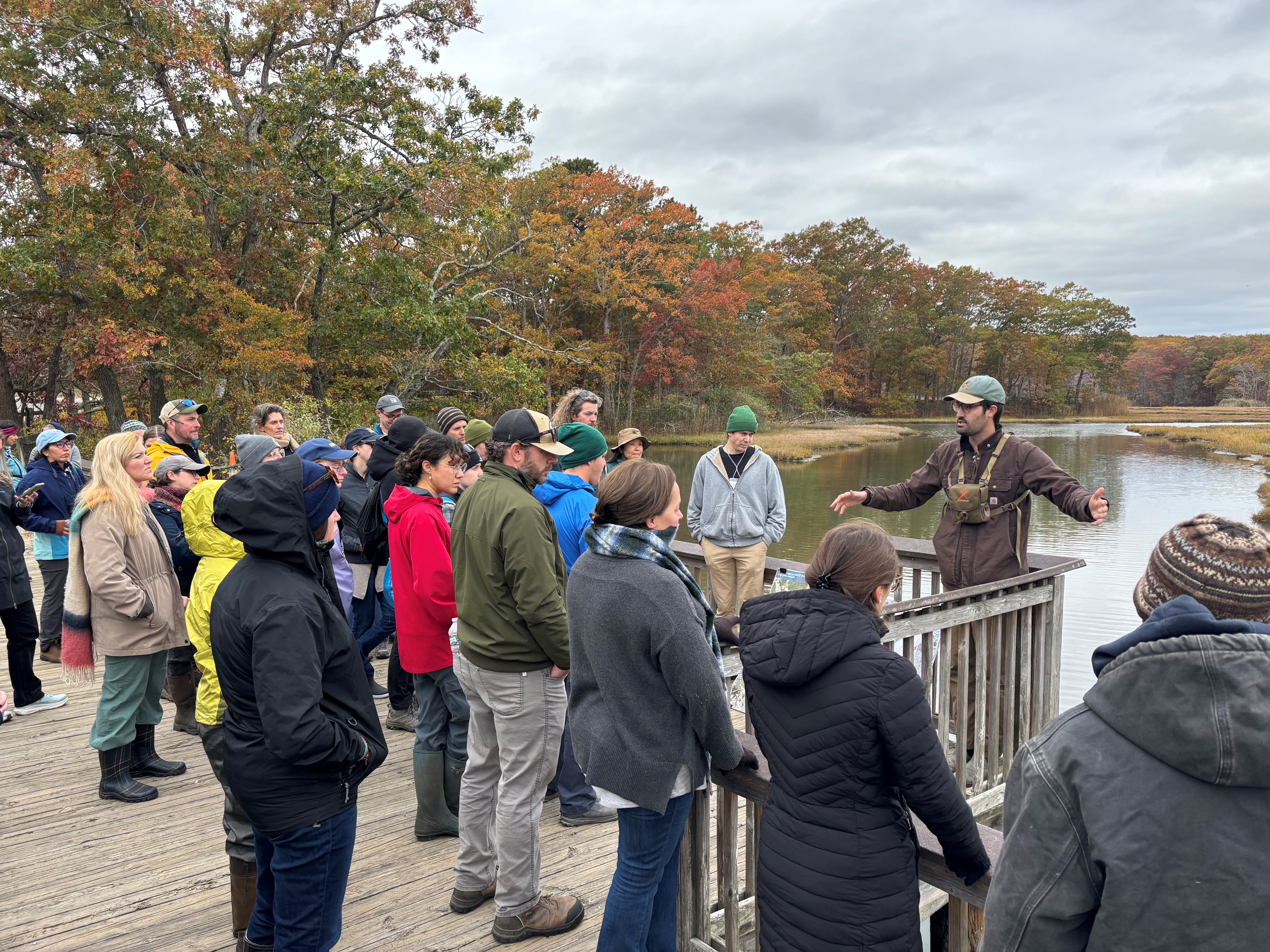 A group of people listening to a TNC staffperson at an outdoor educational event beside a scenic marsh lake surrounded by autumn foliage.
