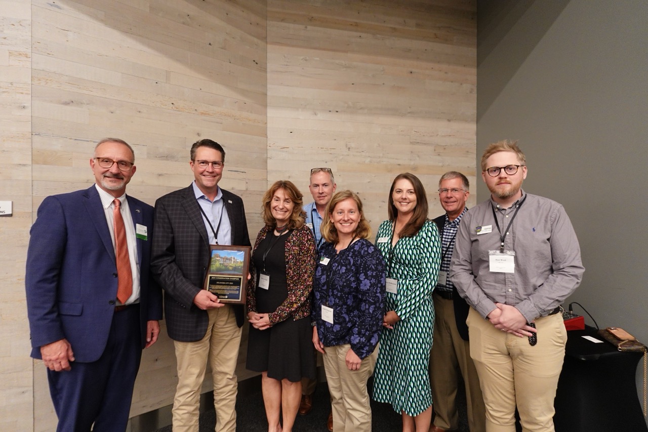 A group of individuals in business attire smiles for a photo with a plaque.