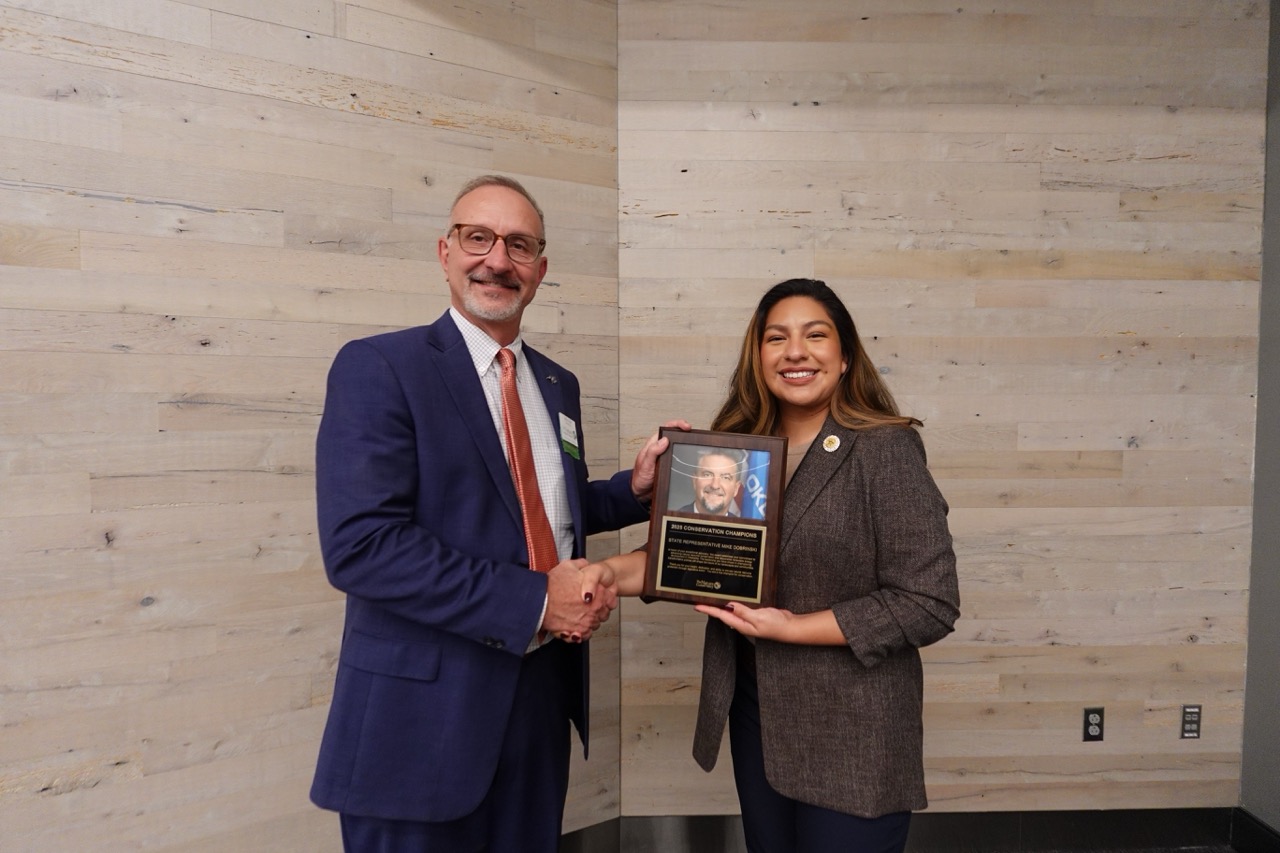 A man and a woman in business attire smile together while holding a plaque.