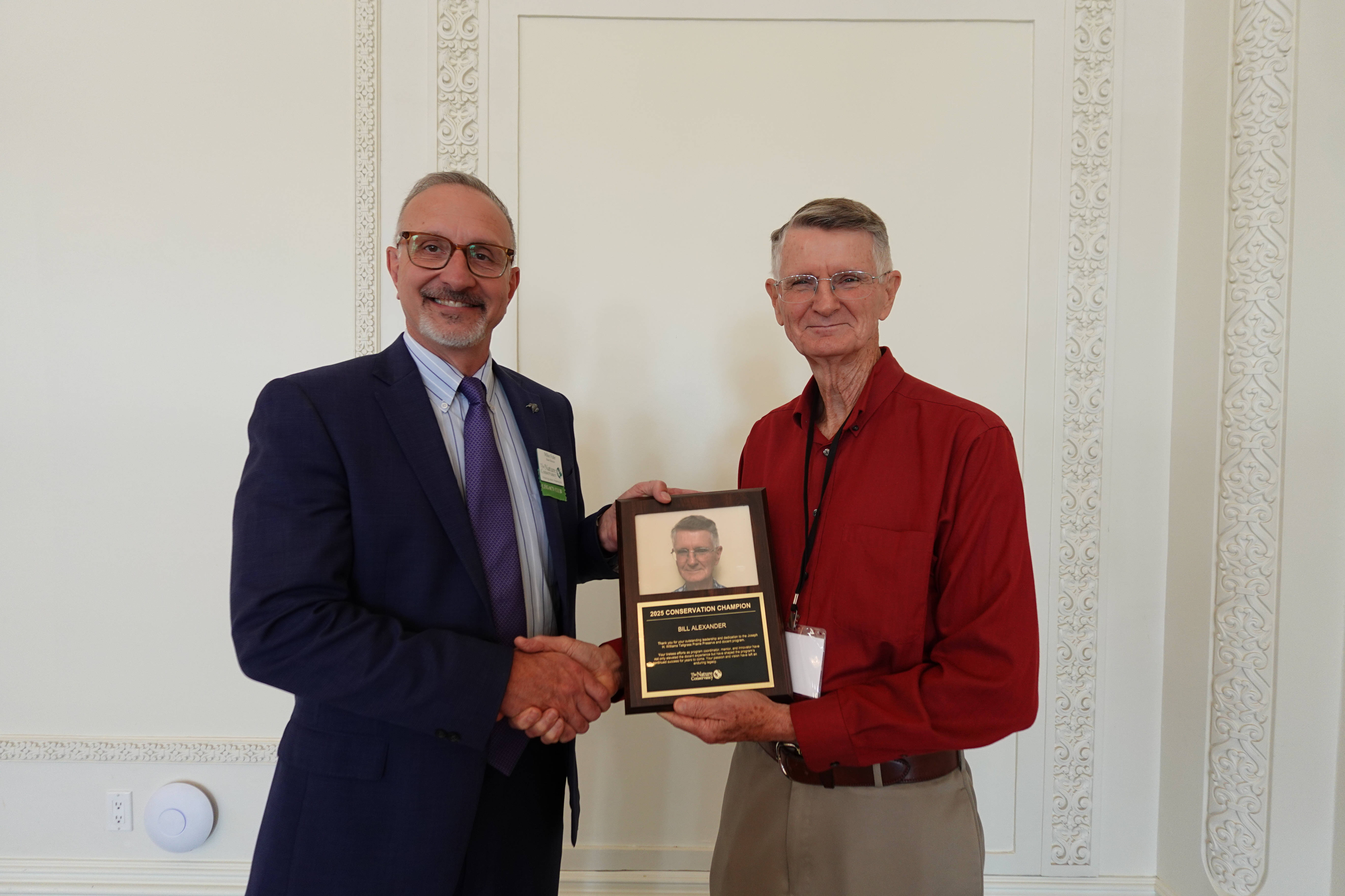 Two men in business attire smile for a photo with a plaque.