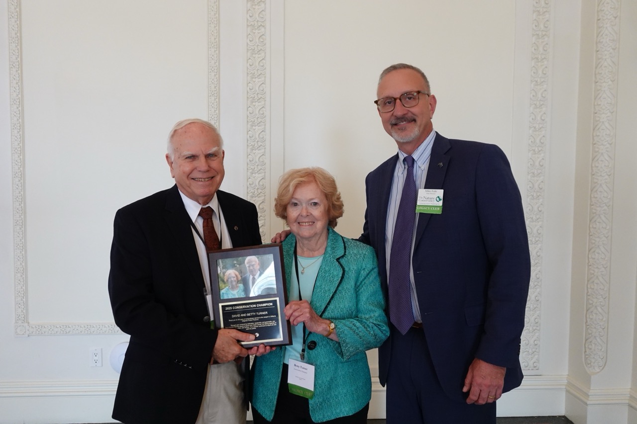 A woman and two men in business attire smile for a photo with a plaque.