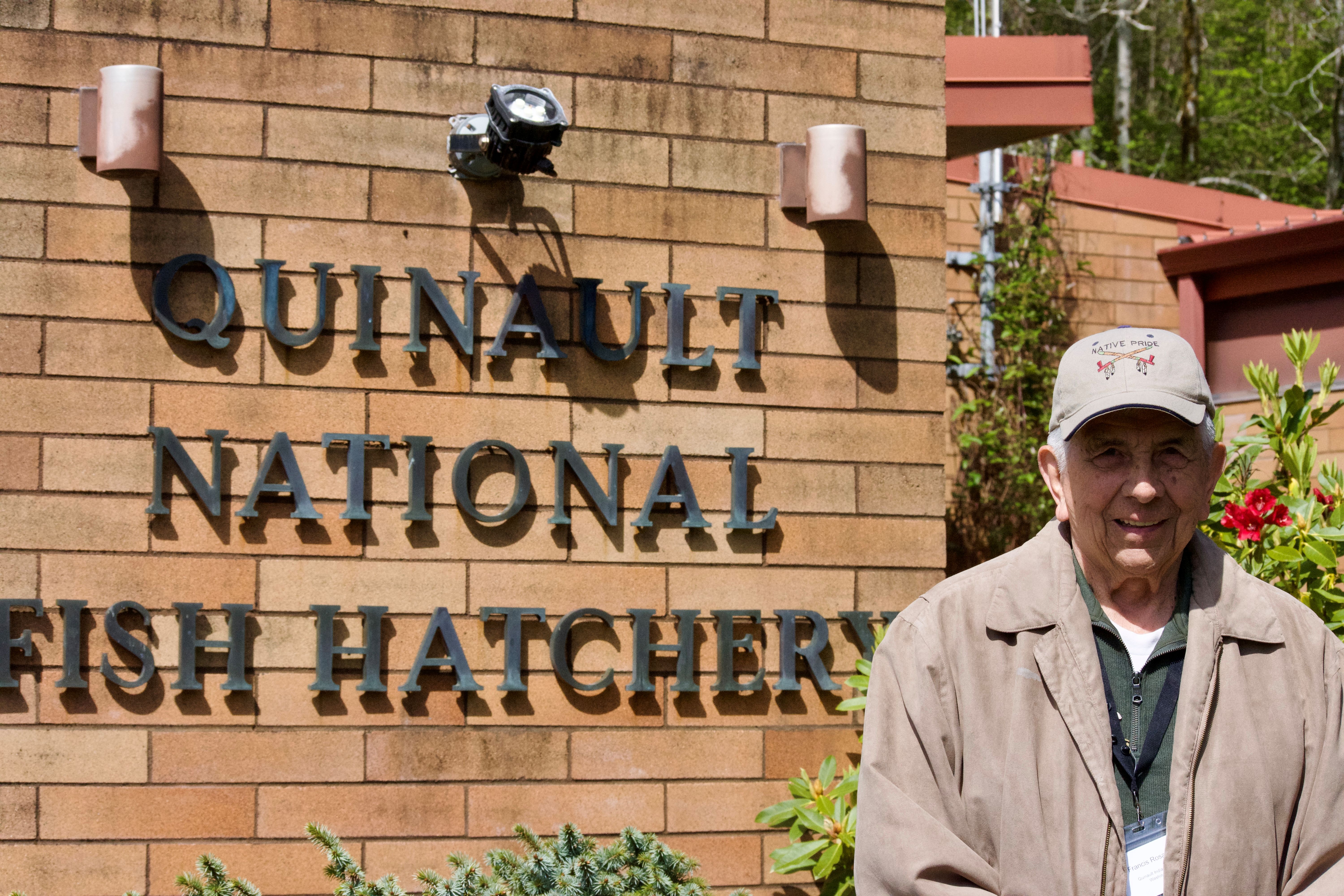 Elder Francis Rosander in front of Quinault National Fish Hatchery signage on the side of a brick building.