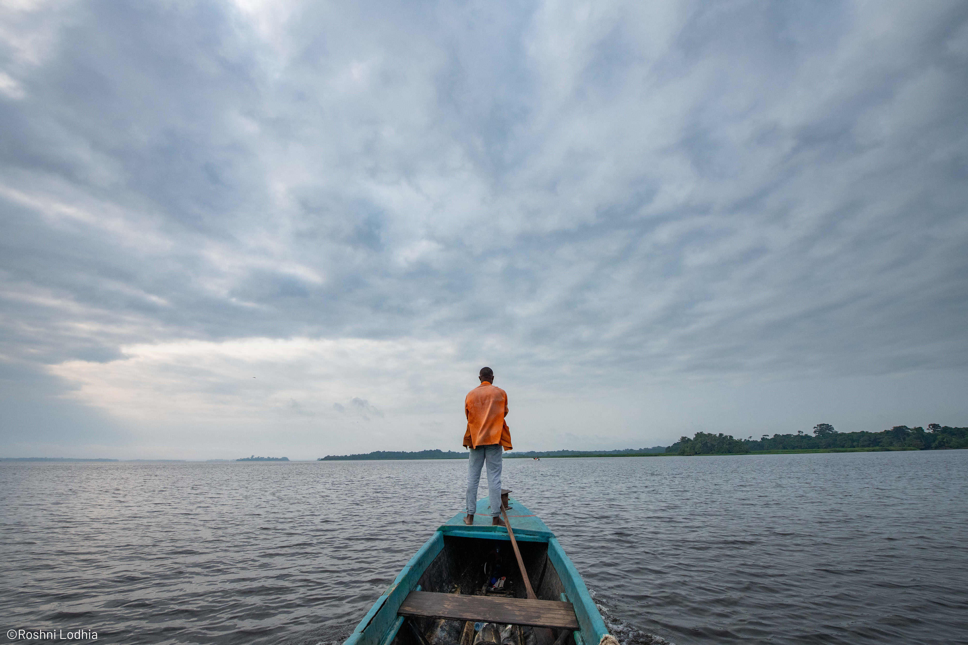 A man in an orange shirt stands at the helm of a small blue fishing boat on a large lake. The sky above is overcast, and a distant forest is visible on the horizon. 
