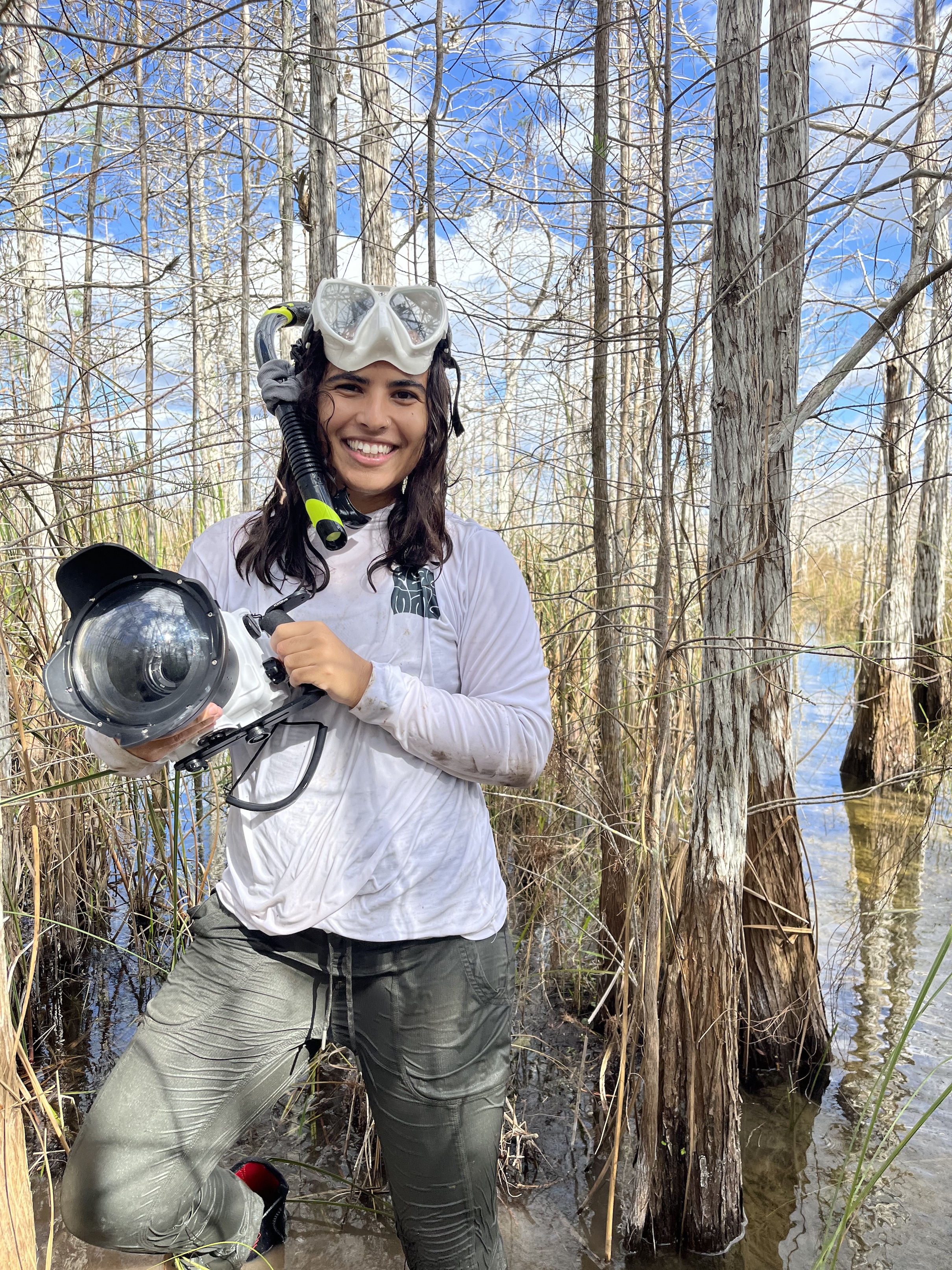 A person smiles while holding a large underwater camera and wearing snorkeling equipment.