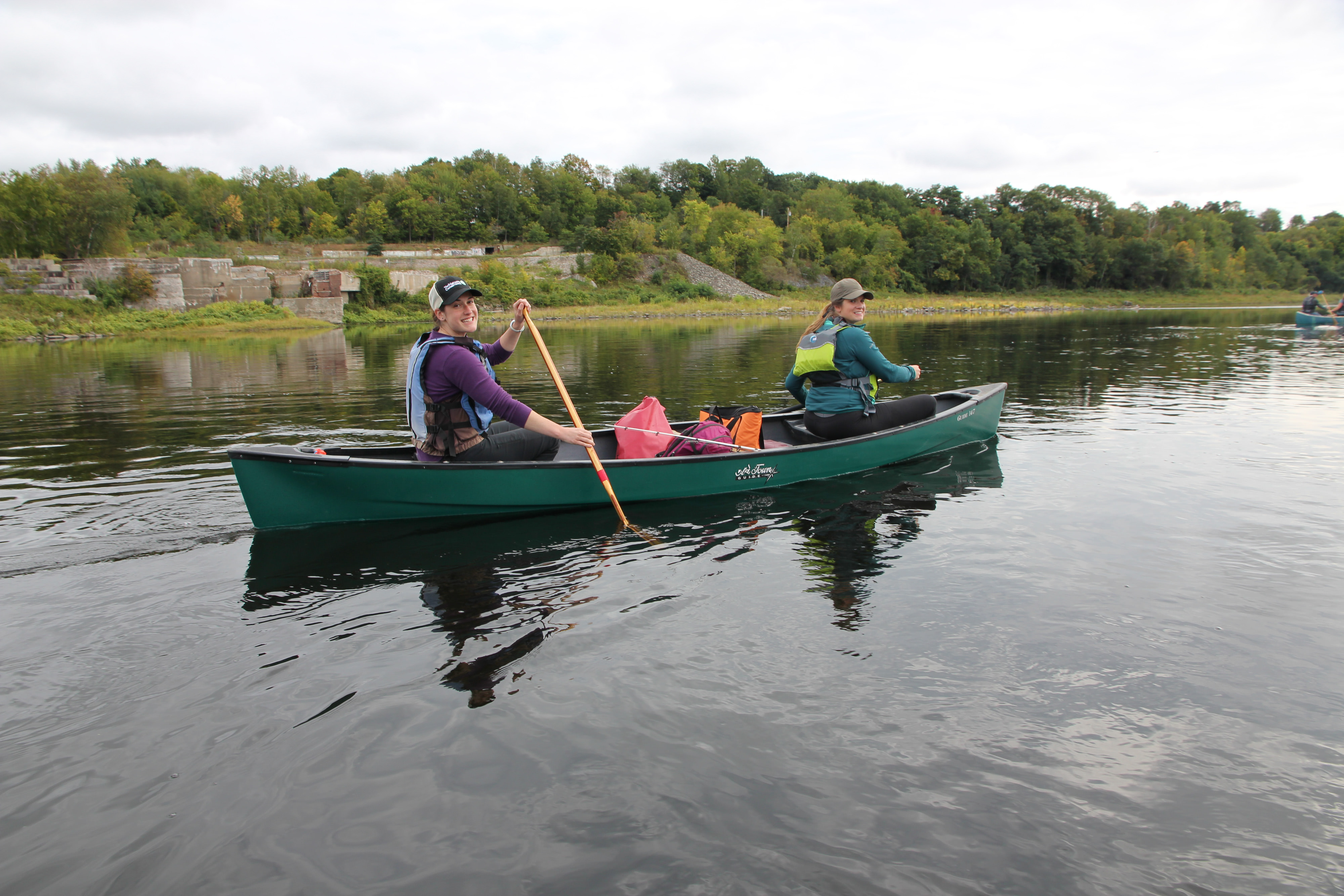 Two women paddle down a river in a green canoe. One woman is wearing a purple pullover, blue life vest, and black jeans with a TNC-branded baseball cap. The other woman is wearing a tan baseball cap, teal jacket, and yellow life vest. 