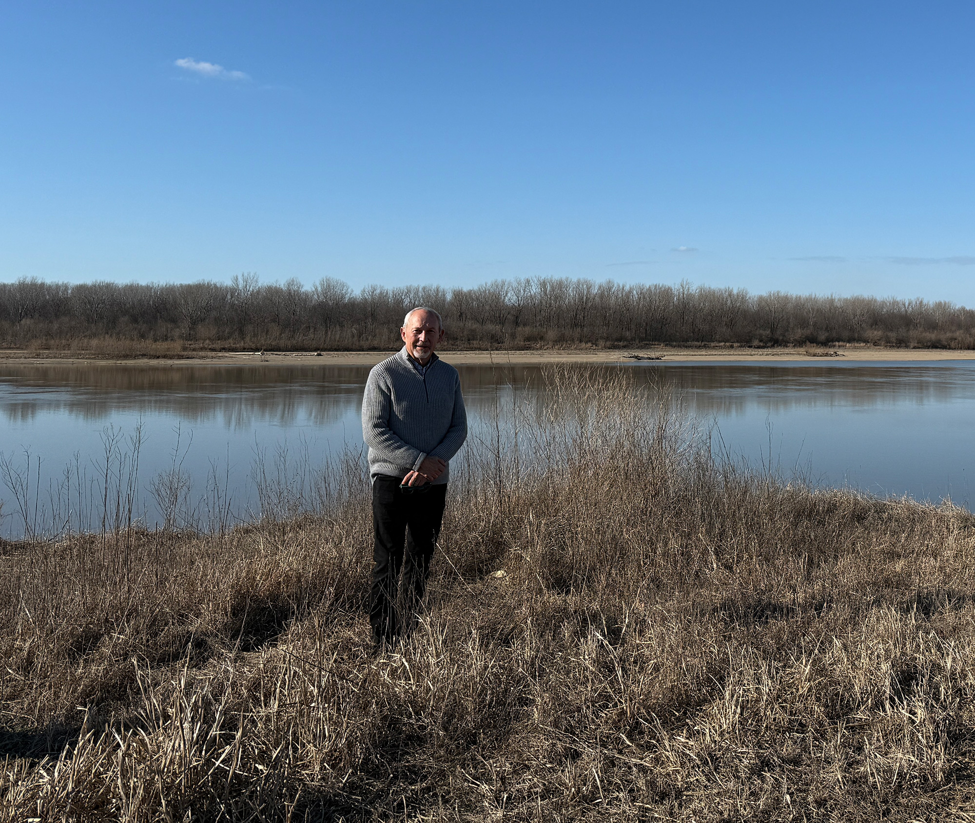 A man stands on the bank of a river amidst dormant brown foliage.