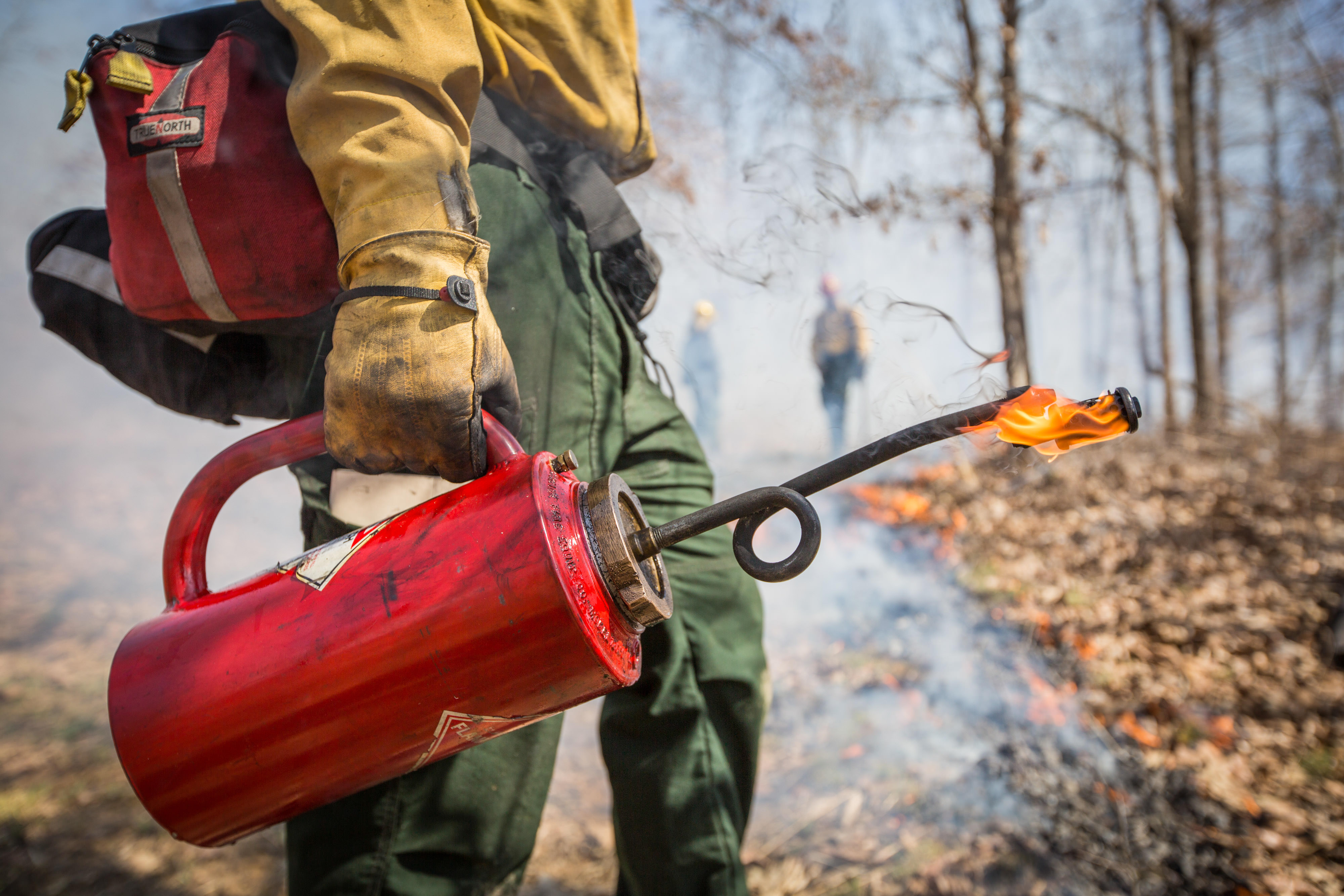A red drip torch apparatus is held by a person wearing gloves and a pack. 