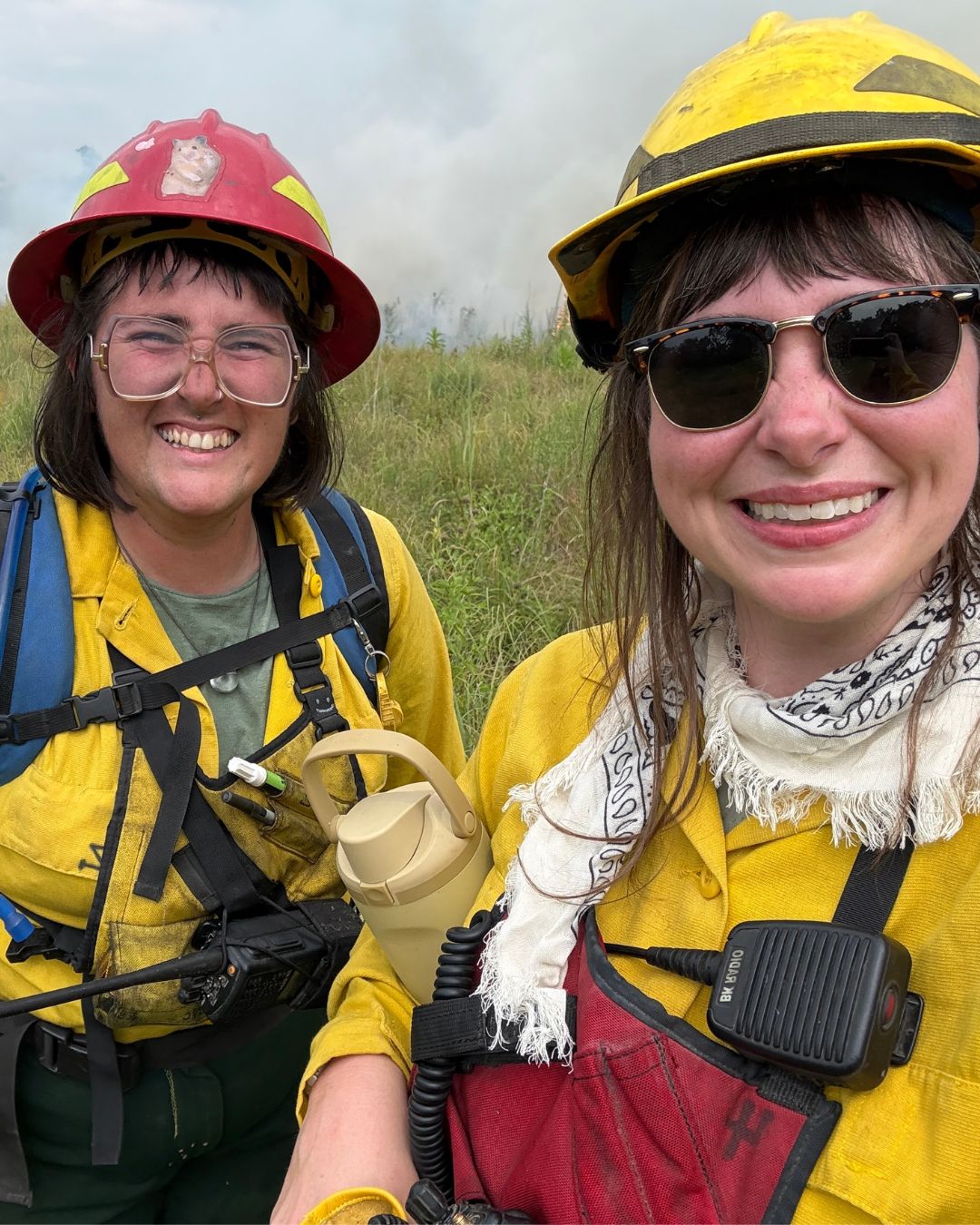 Two women wearing hard hats and bright yellow clothing smile at the camera in their fire gear with smoke rising behind them.