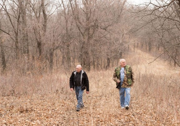 Two men in coats walk along a leaf filled path, bare trees in the background. 