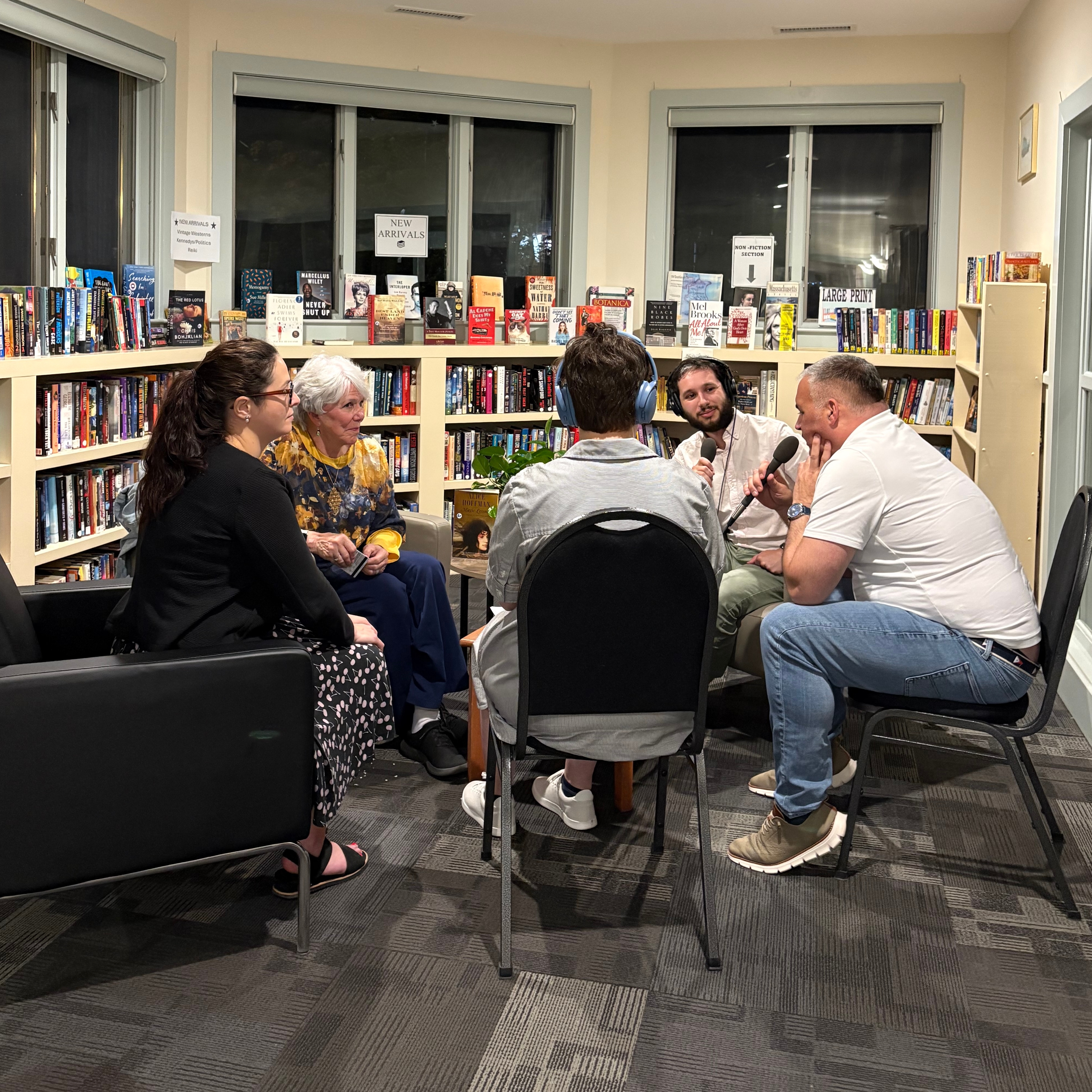 Several people gather in a circle of chairs in a public library, one person wears headphones and holds a recording device.