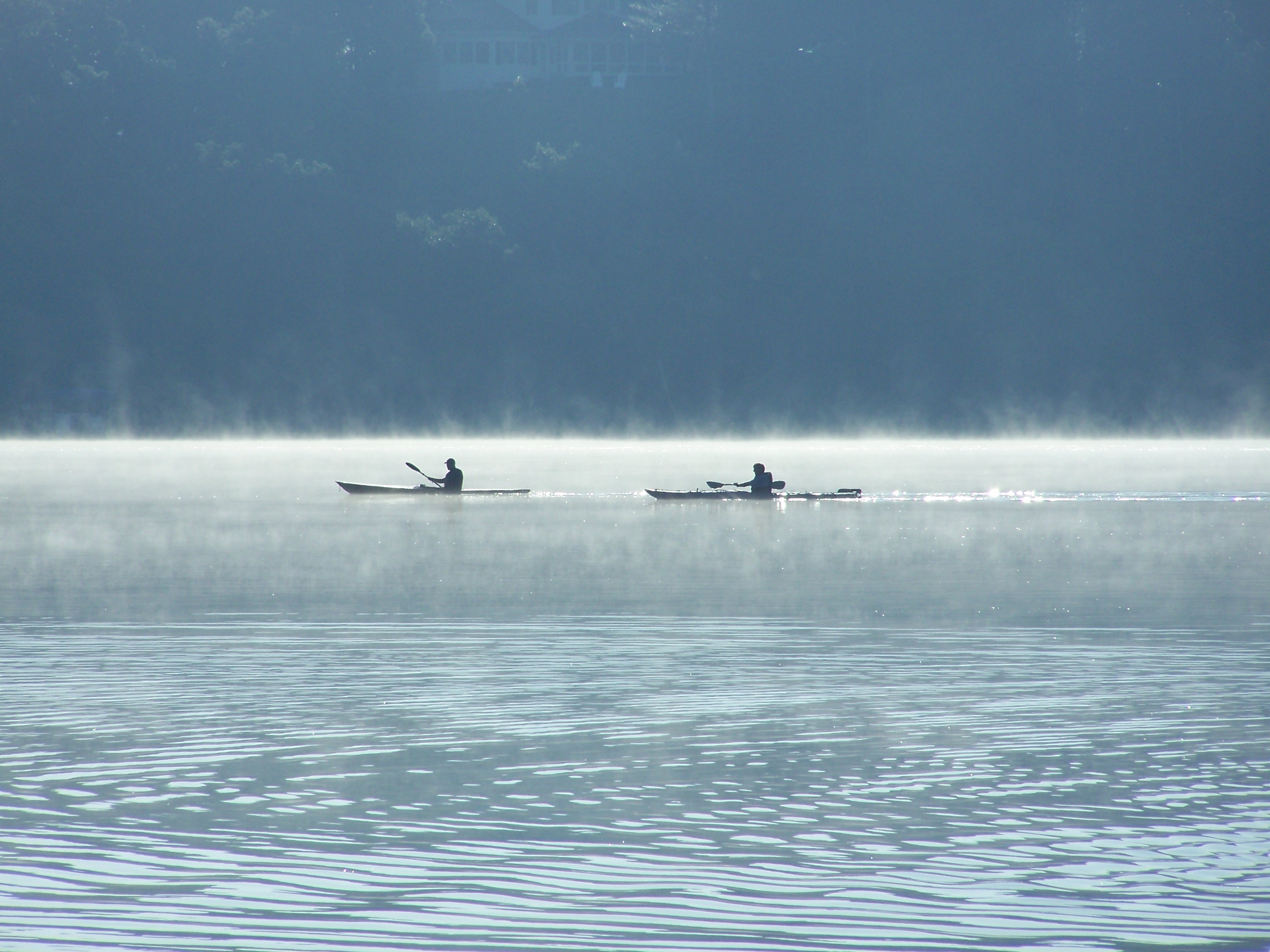 Two kayakers paddle along the morning fog on a tree-lined blue lake. 