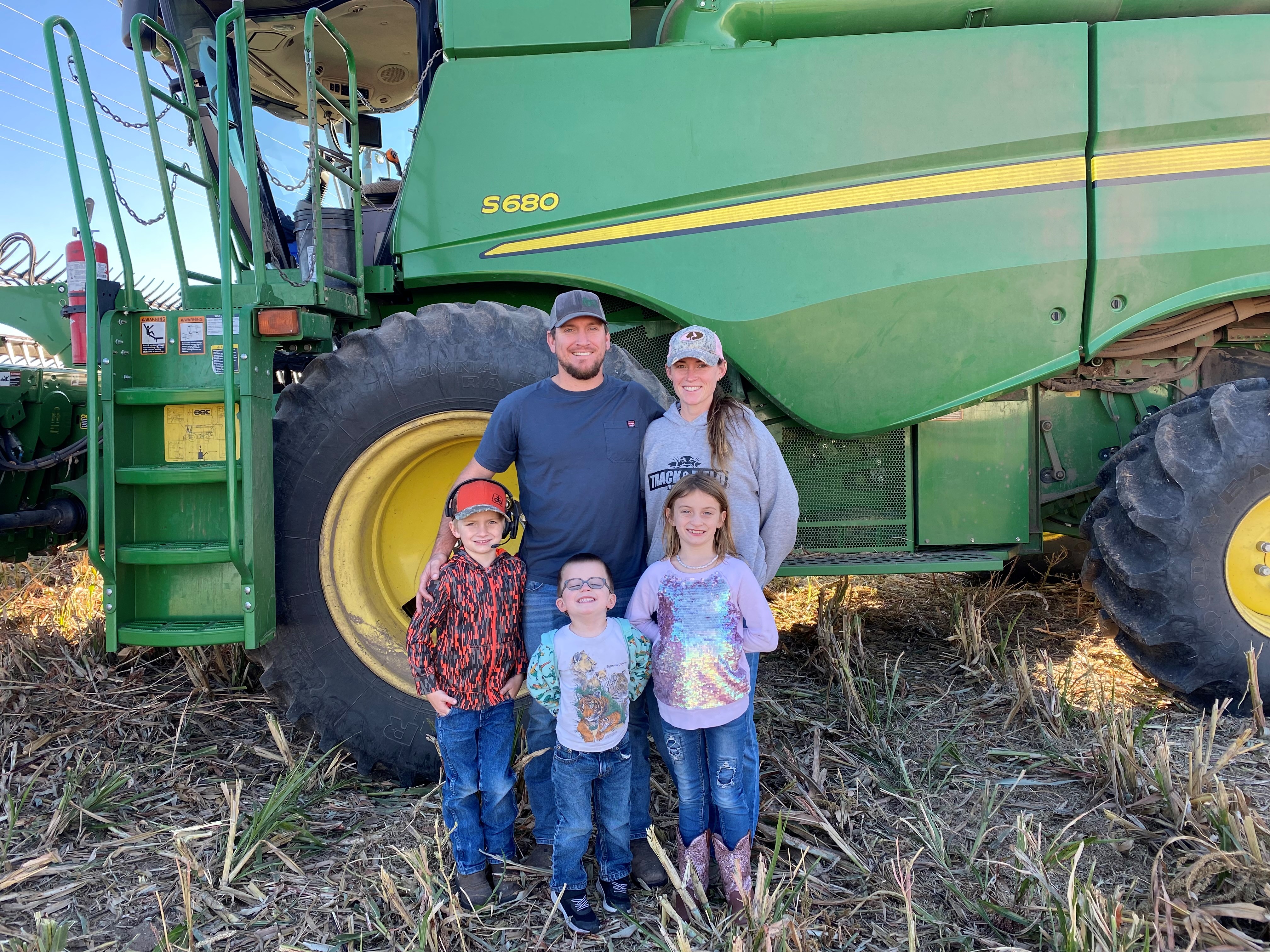 A family with three young children stand in front of a large green piece of farm equipment in the middle of a field.