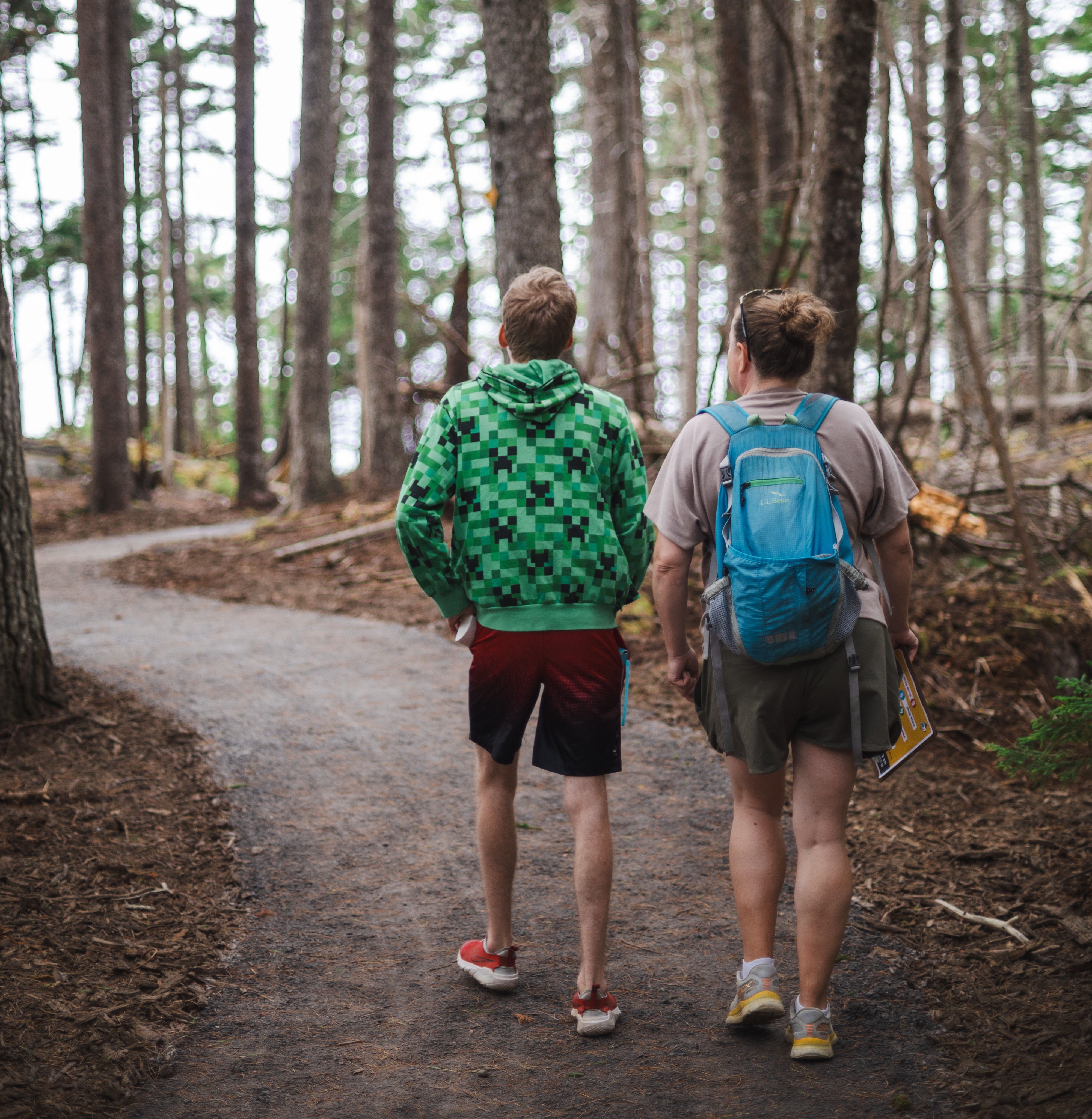 Two individuals, a youth and an adult with a backpack, walk on a crushed gravel trail out toward a coastal view.