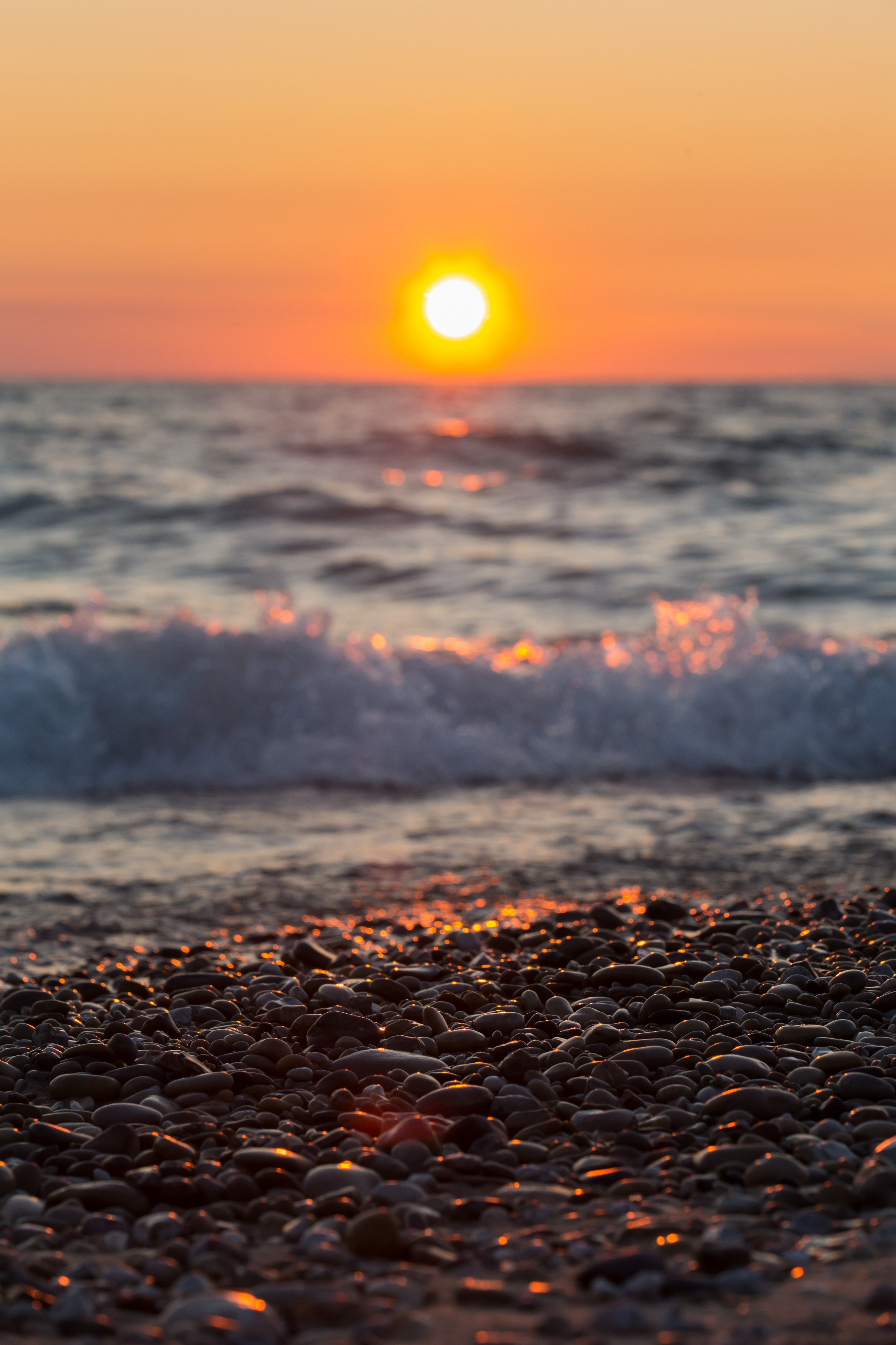 A bright orange sunset on Lake Michigan, with a closeup view of beach pebbles in the foreground as a wave rolls in.