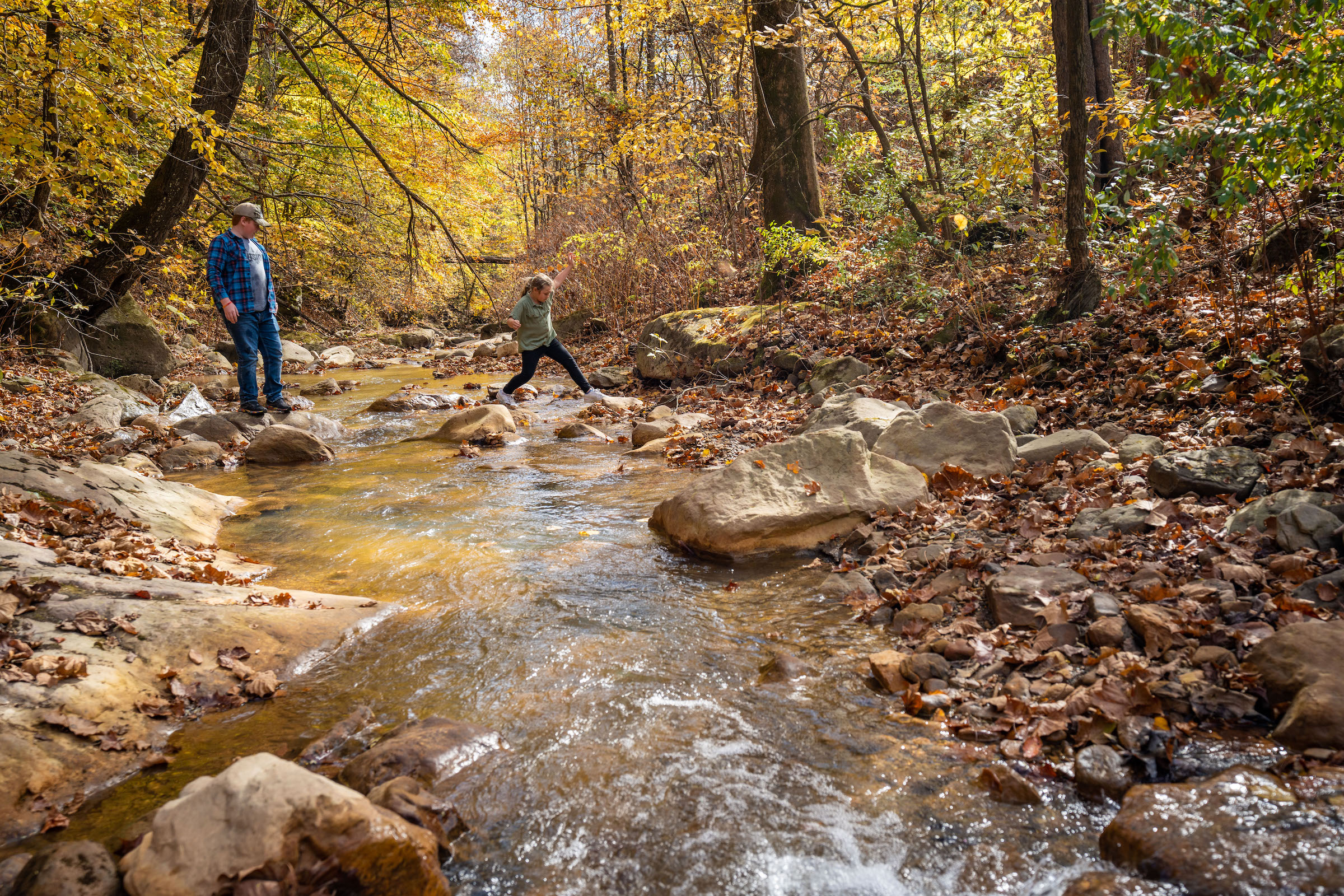 Child leaping across a stream with guardian looking on.