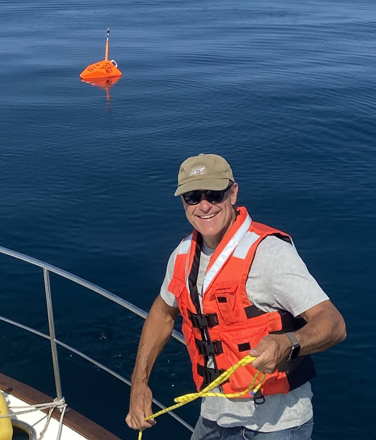 Matt Merrifield wears a hat and lifejacket and smiles on a boat with a Scout buoy in the water behind him. 