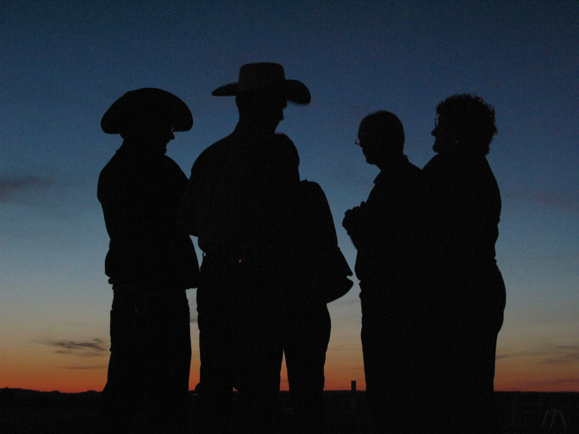 Several people are silhouetted against a dusk sky with a low-setting sun casting an orange glow on the horizon.