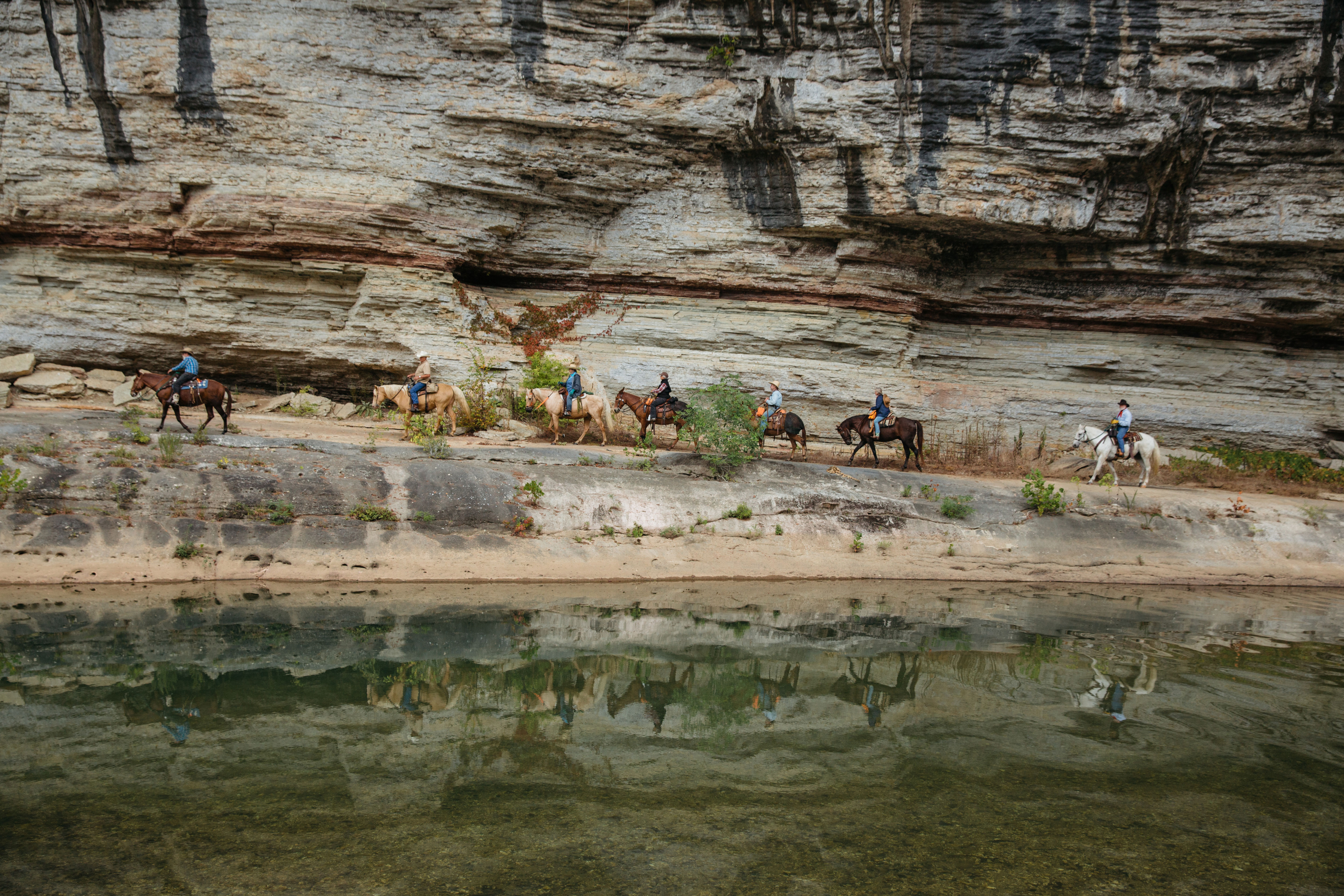 A line of horses and riders move up cliffs above water.