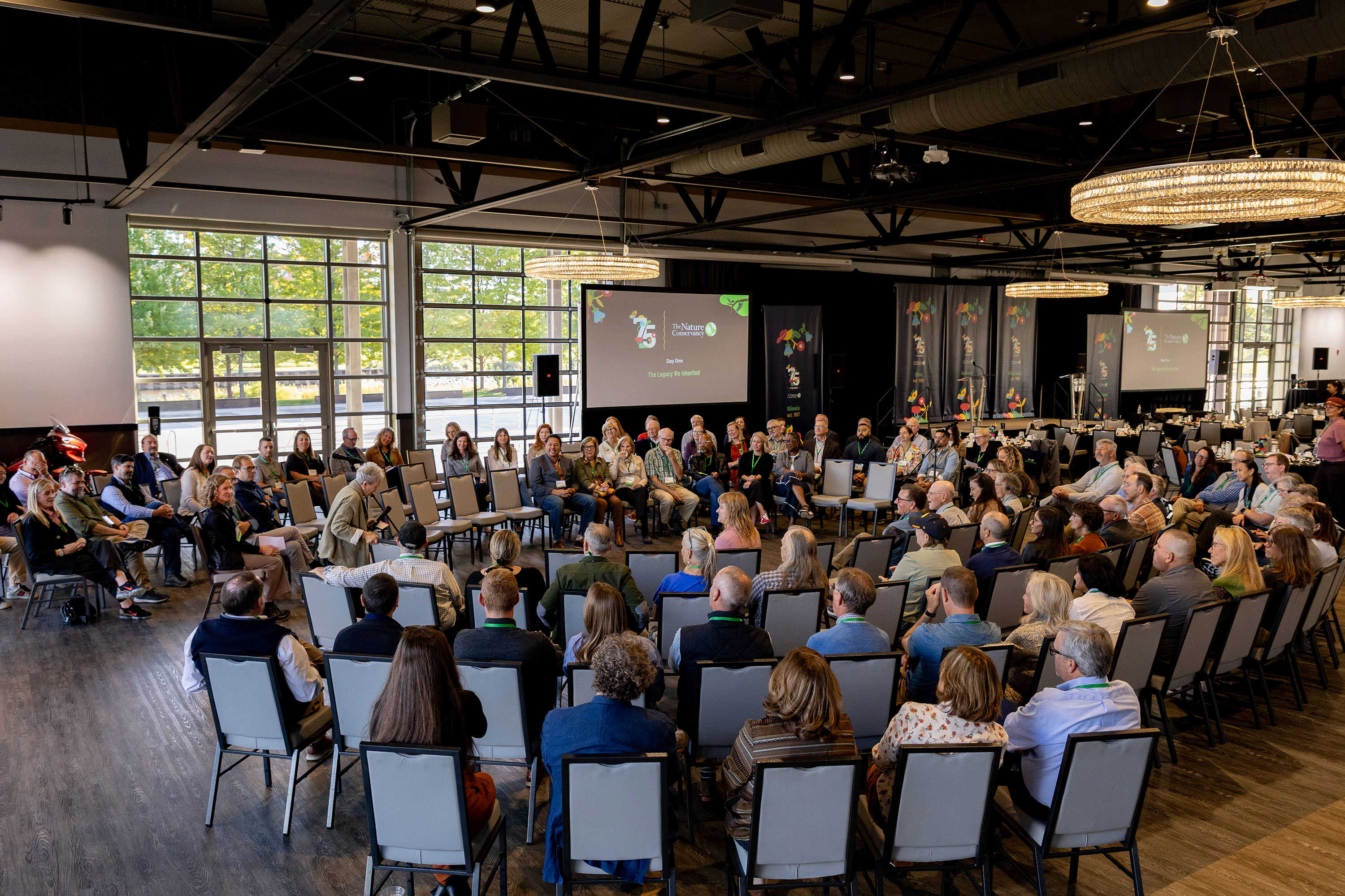 A group of people seated in chairs in a big room face a screen and row of large windows.