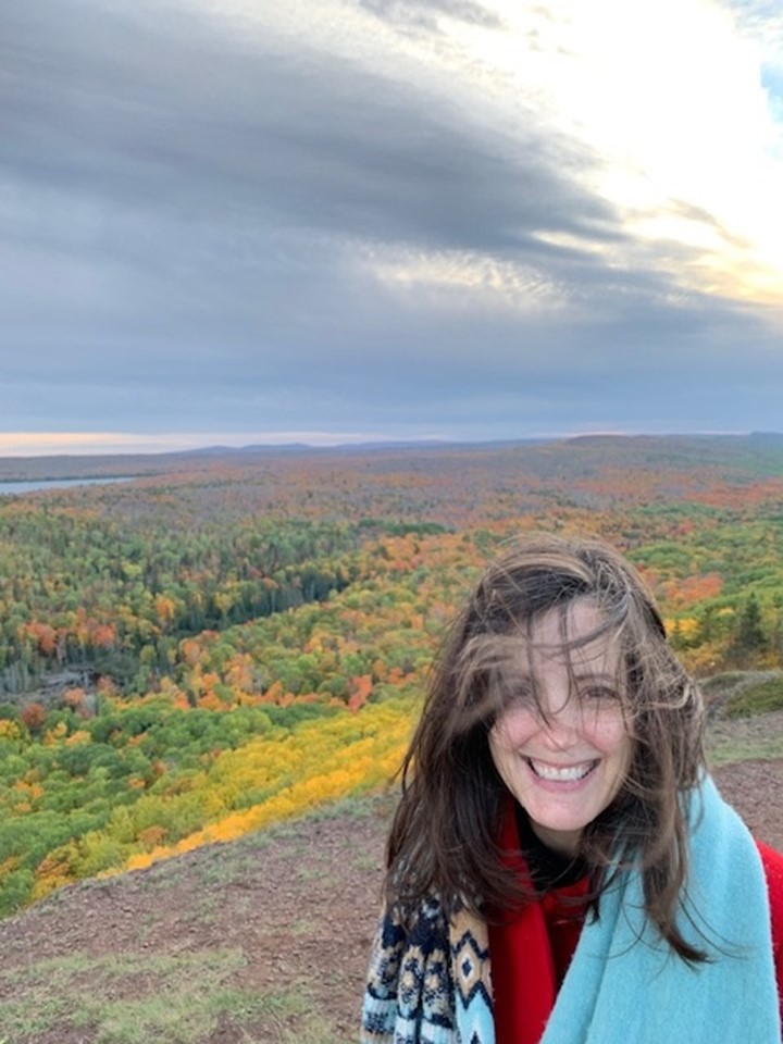 A woman in standing outside in front of a fall forest. 