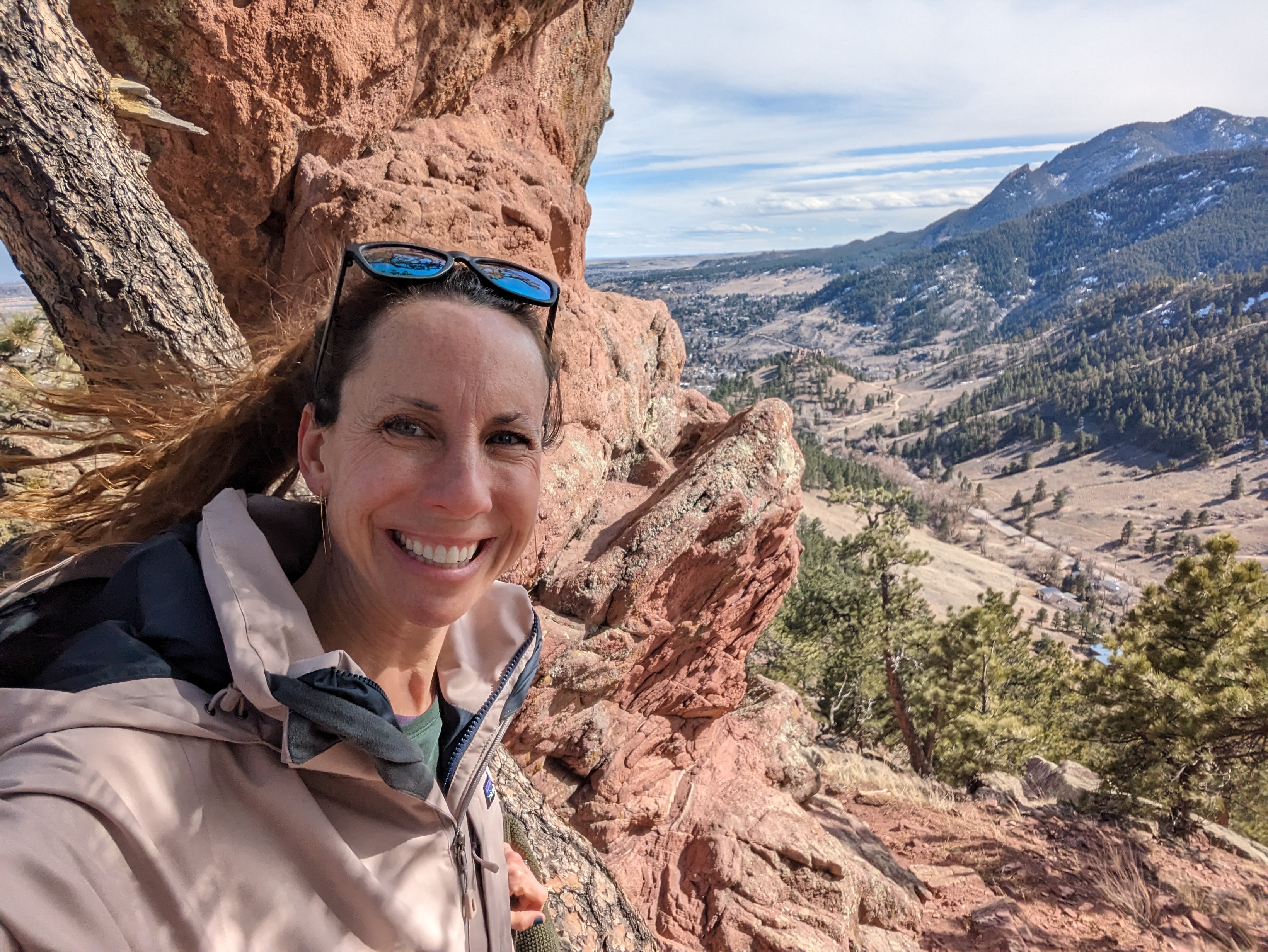 A woman with long hair smiling at the camera as she stands among red rocks and mountains. 