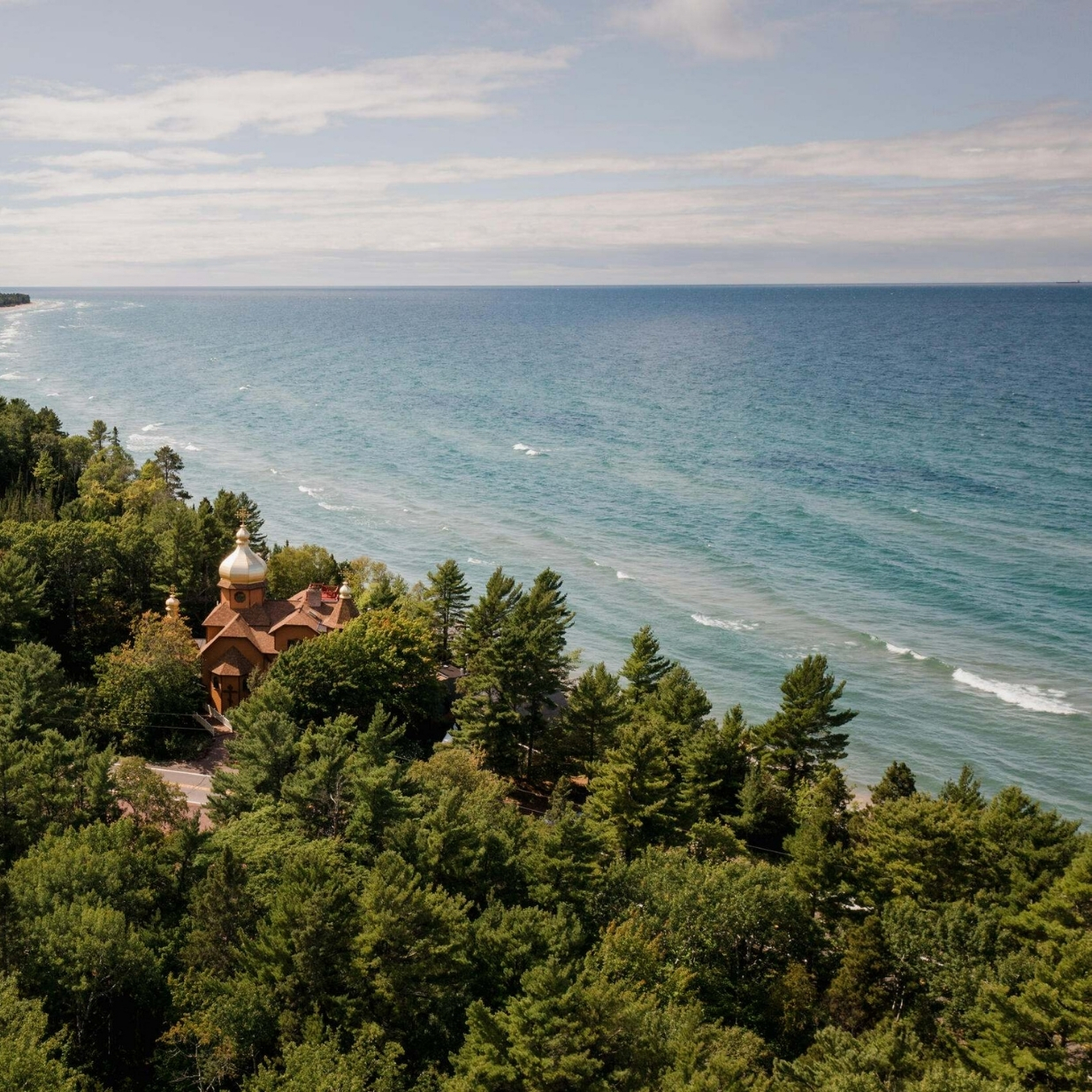 An aerial view of a forested shoreline beside a large blue lake, with a small wooden chapel featuring gold-colored domes nestled among the trees. Gentle waves line the coast beneath a partly cloudy sky.