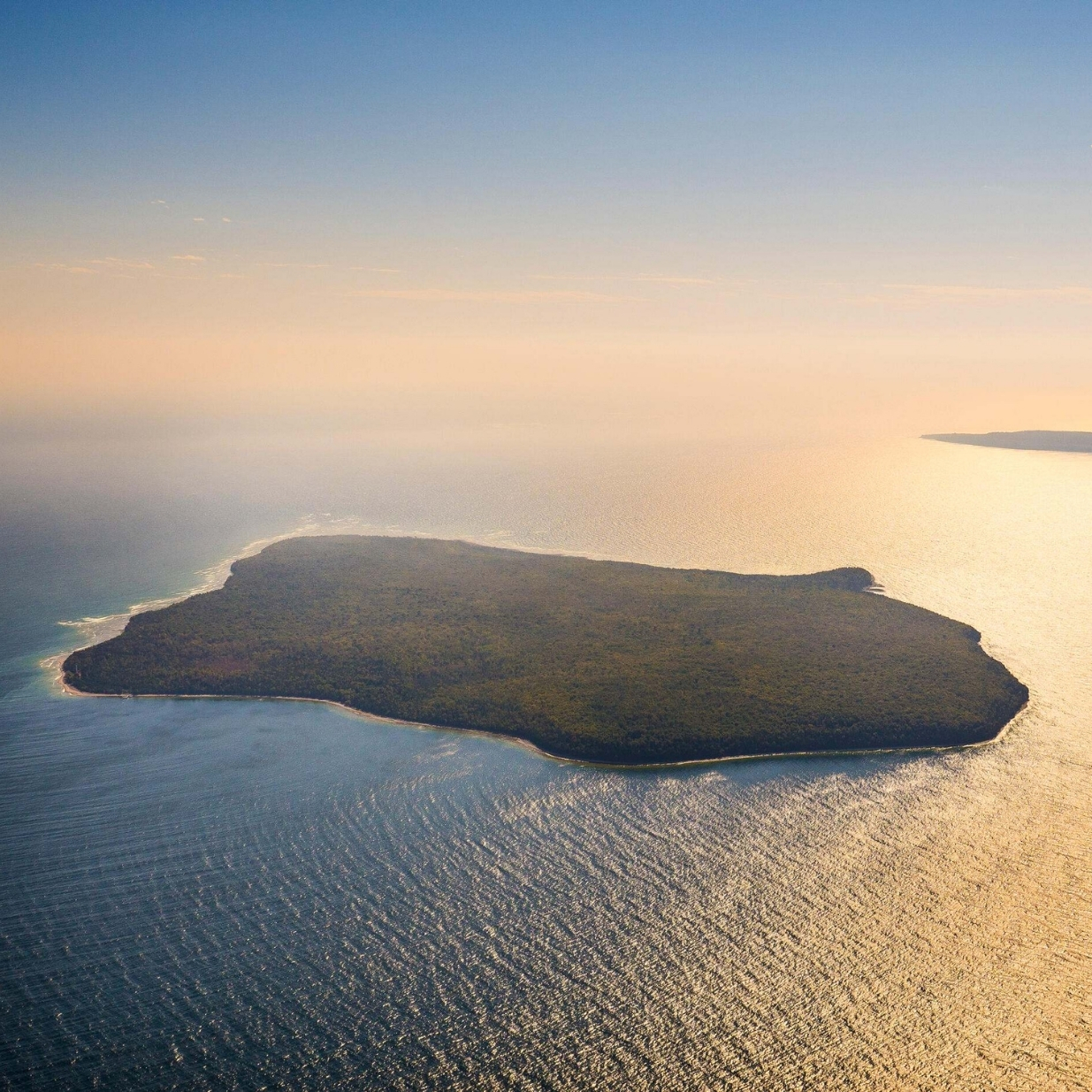 Aerial view of a forest-covered island surrounded by shimmering blue and gold water, with sunlight reflecting across the lake and a hazy horizon beneath a clear sky.