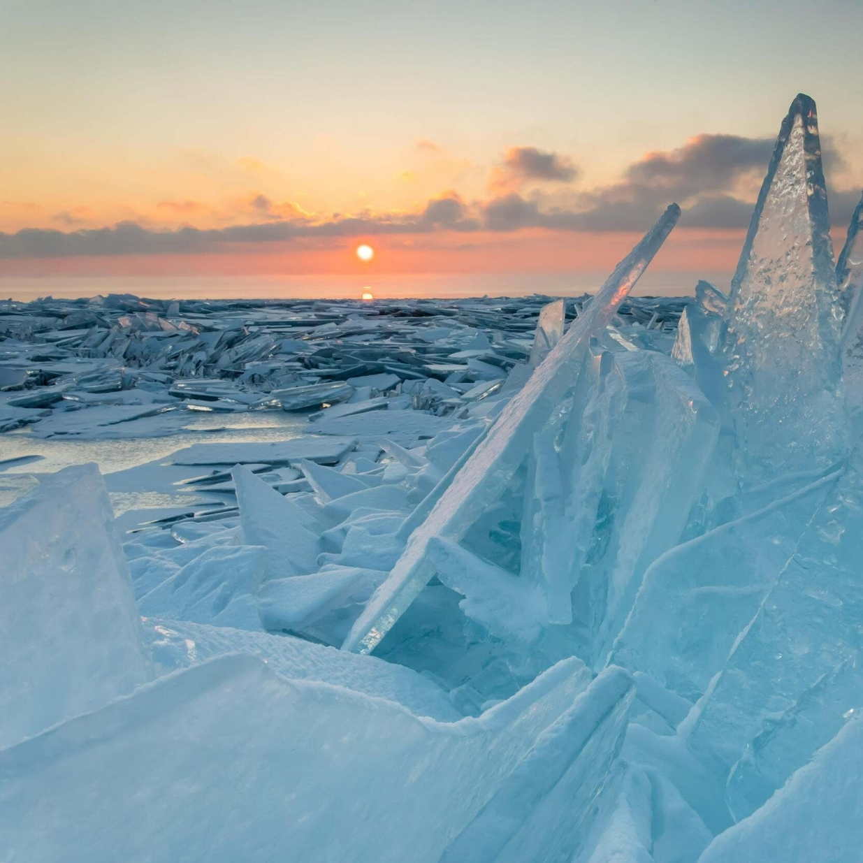 Jagged blue ice formations rise from a frozen lakeshore at sunrise, with broken ice sheets stretching into the distance beneath a glowing orange sun and pastel-colored sky.